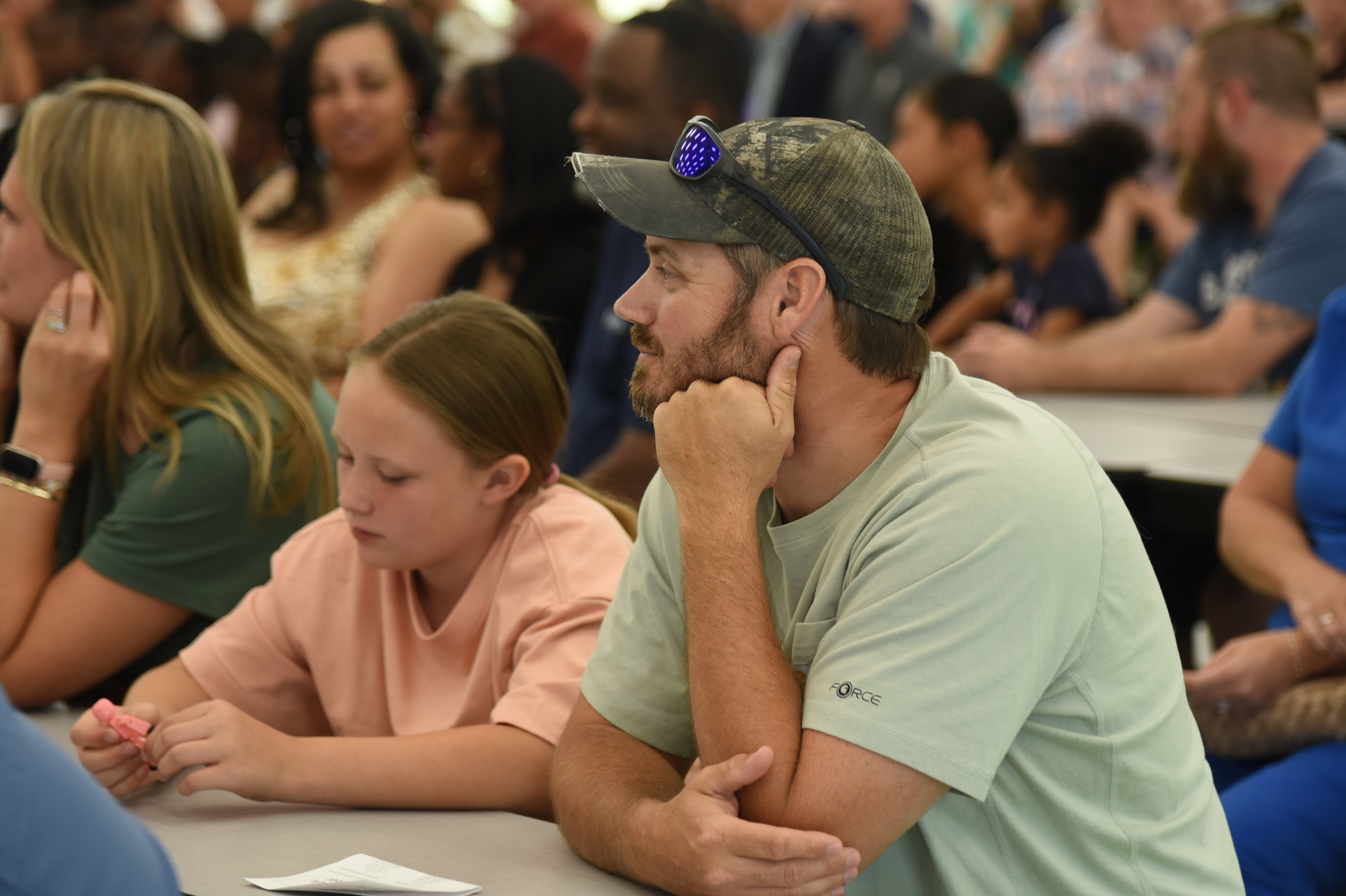 People sit at tables in a meeting. A man rests his chin on his hand, while a girl looks down.
