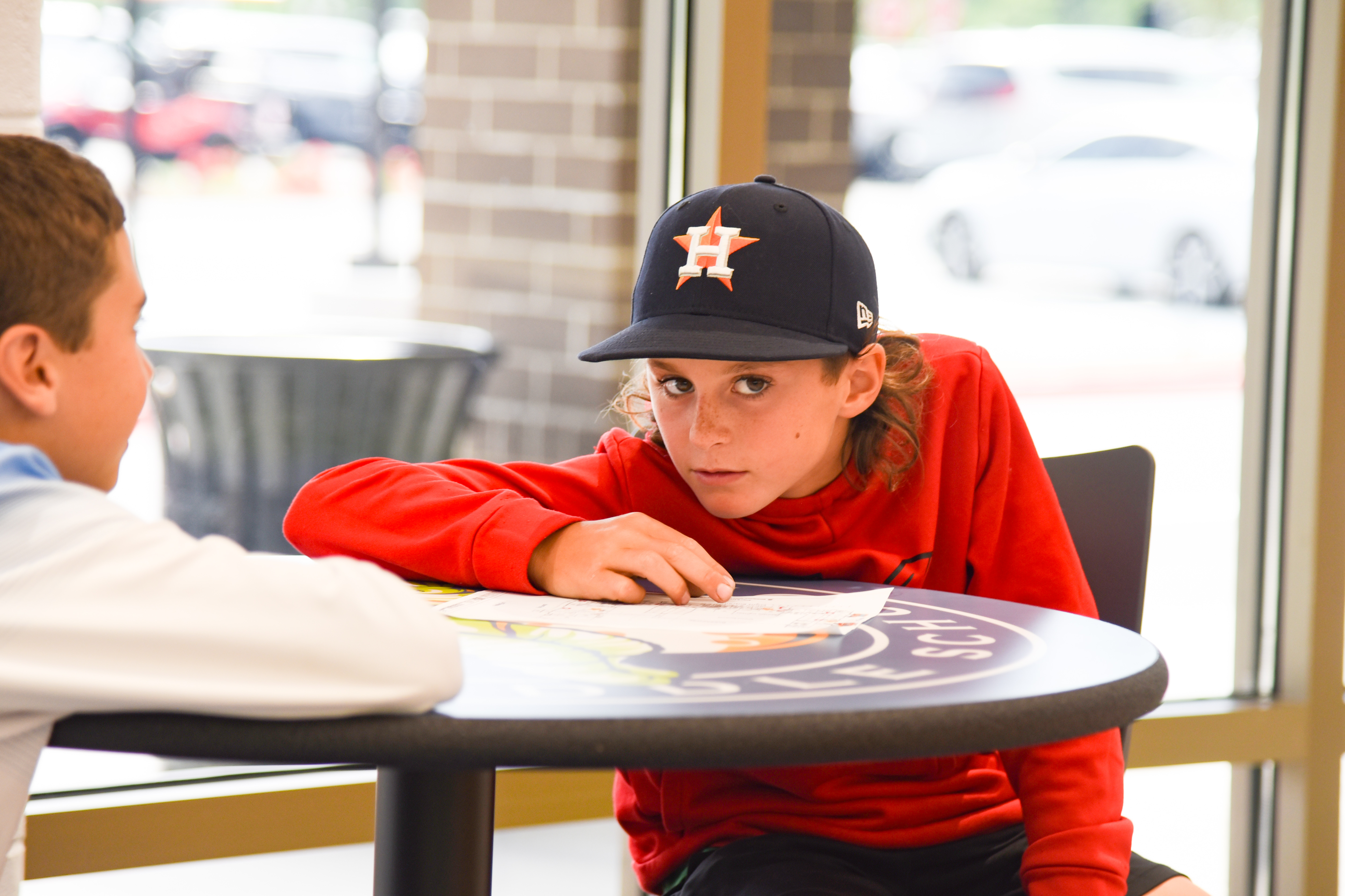 Two boys sit at a table; one looks at paper while the other listens. They wear casual clothing.