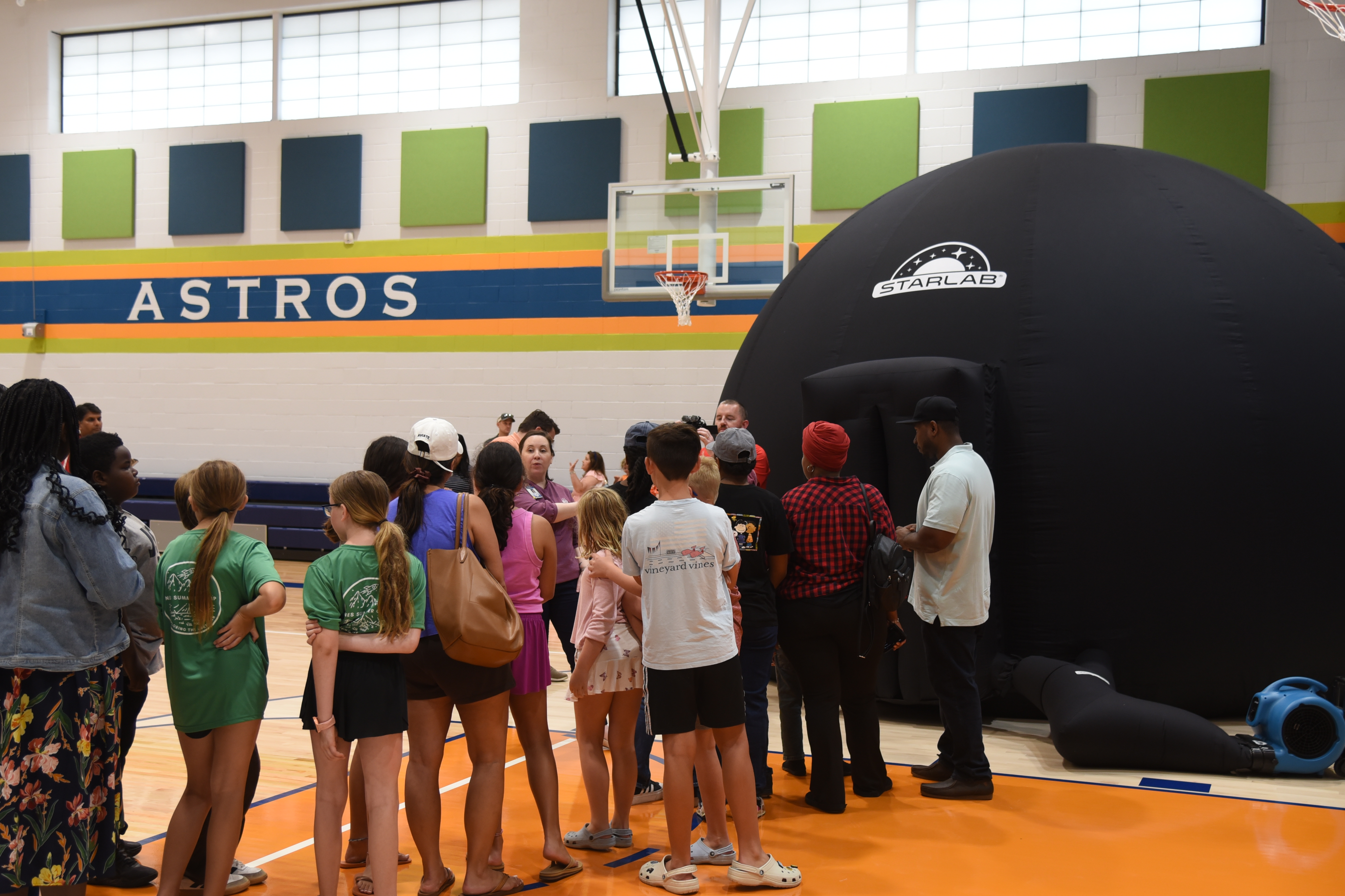 A group of people gather around an inflatable dome inside a basketball court with "Astros" on the wall.