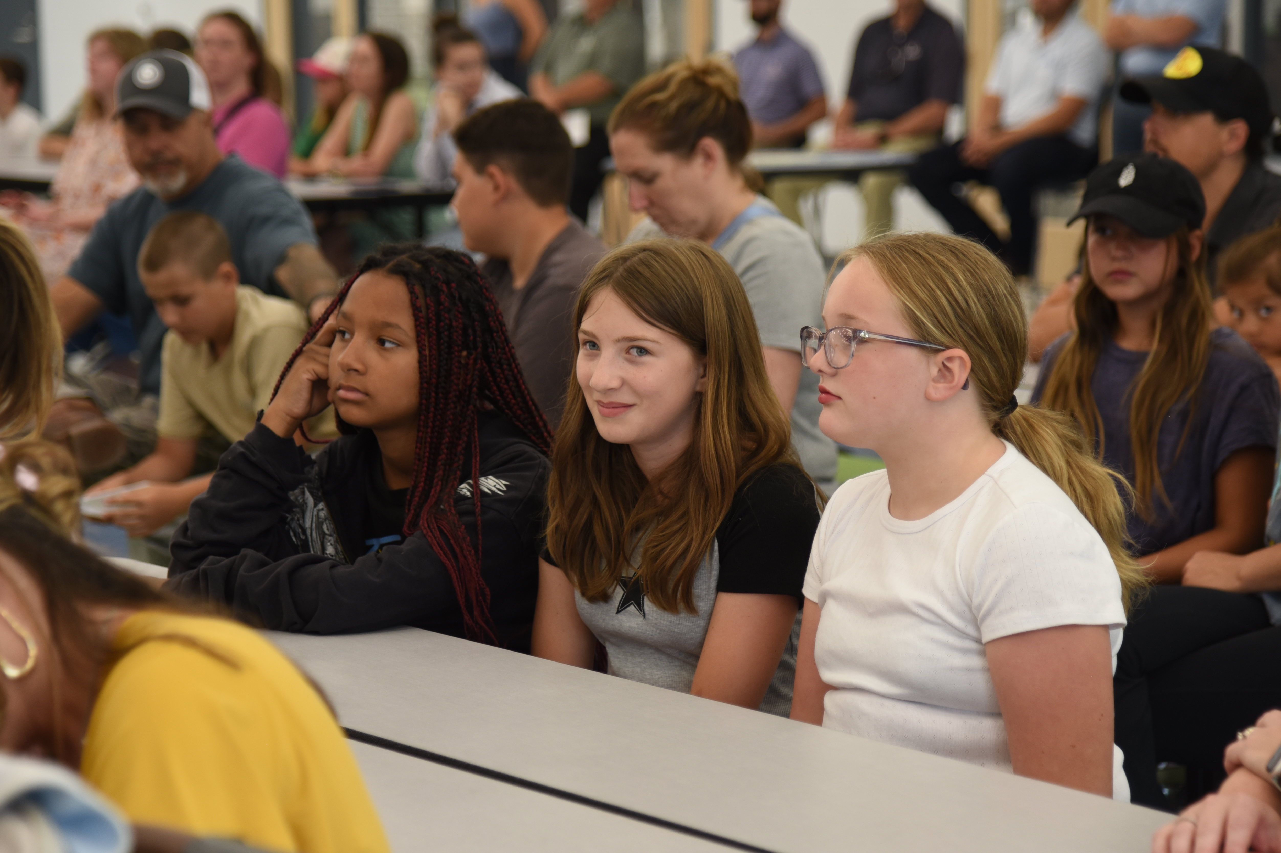 Multiple individuals, including students, seated at tables, looking towards the front of a room.