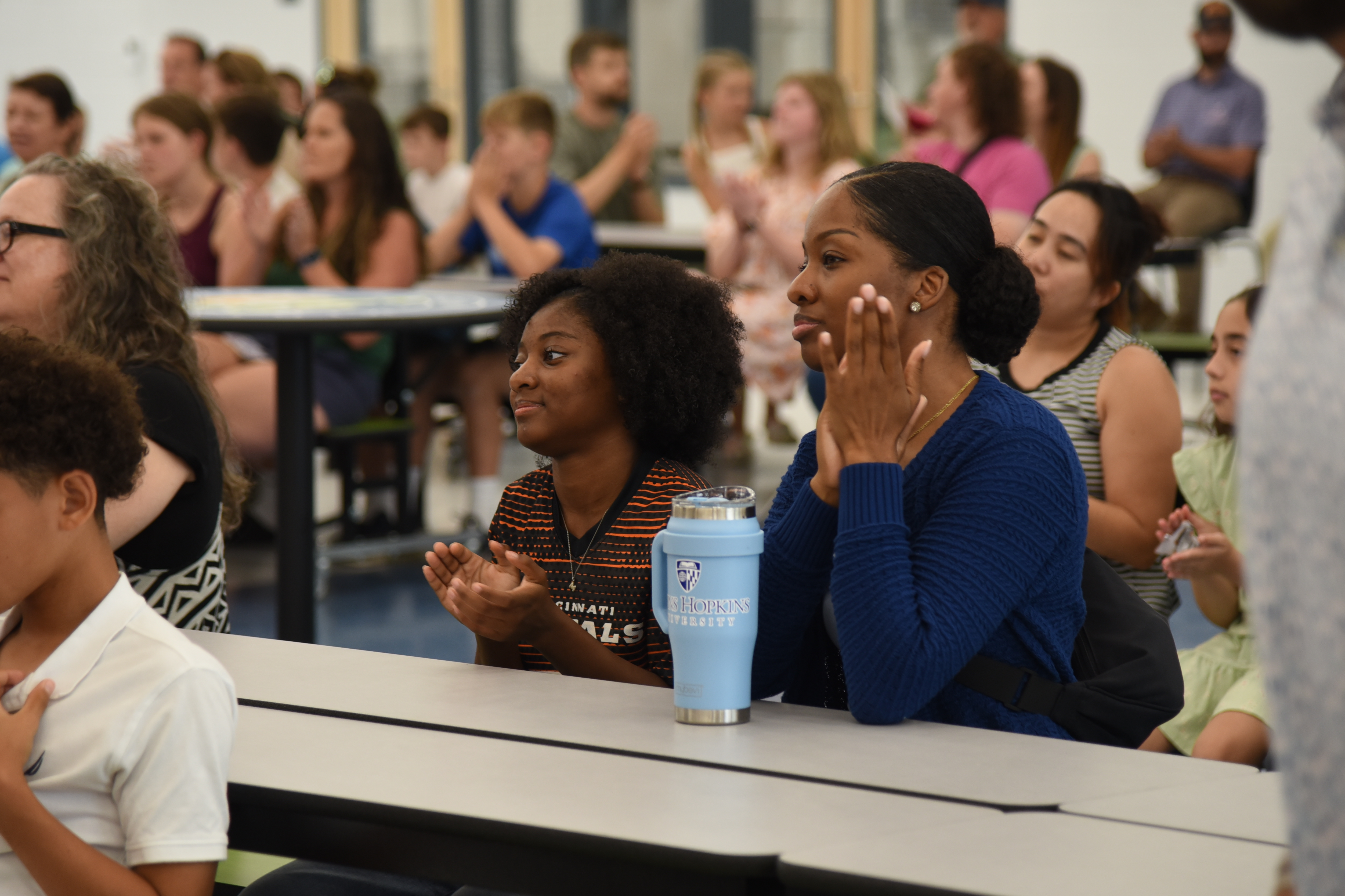 A group of people sit at tables in a room, clapping. A woman in blue claps with her hands together.