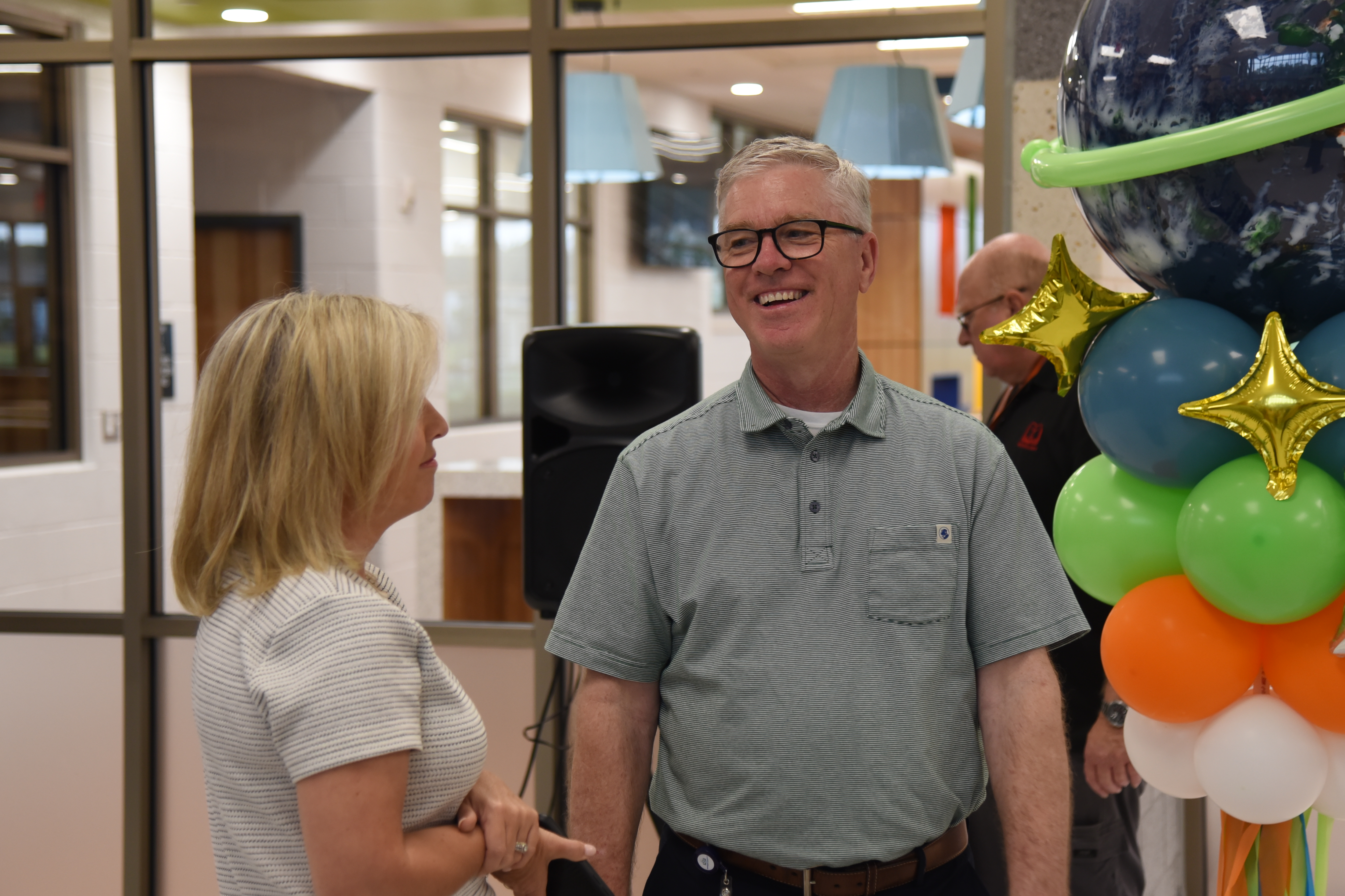 A man and woman stand together near a large balloon display, both smiling. The man wears glasses and a belt.