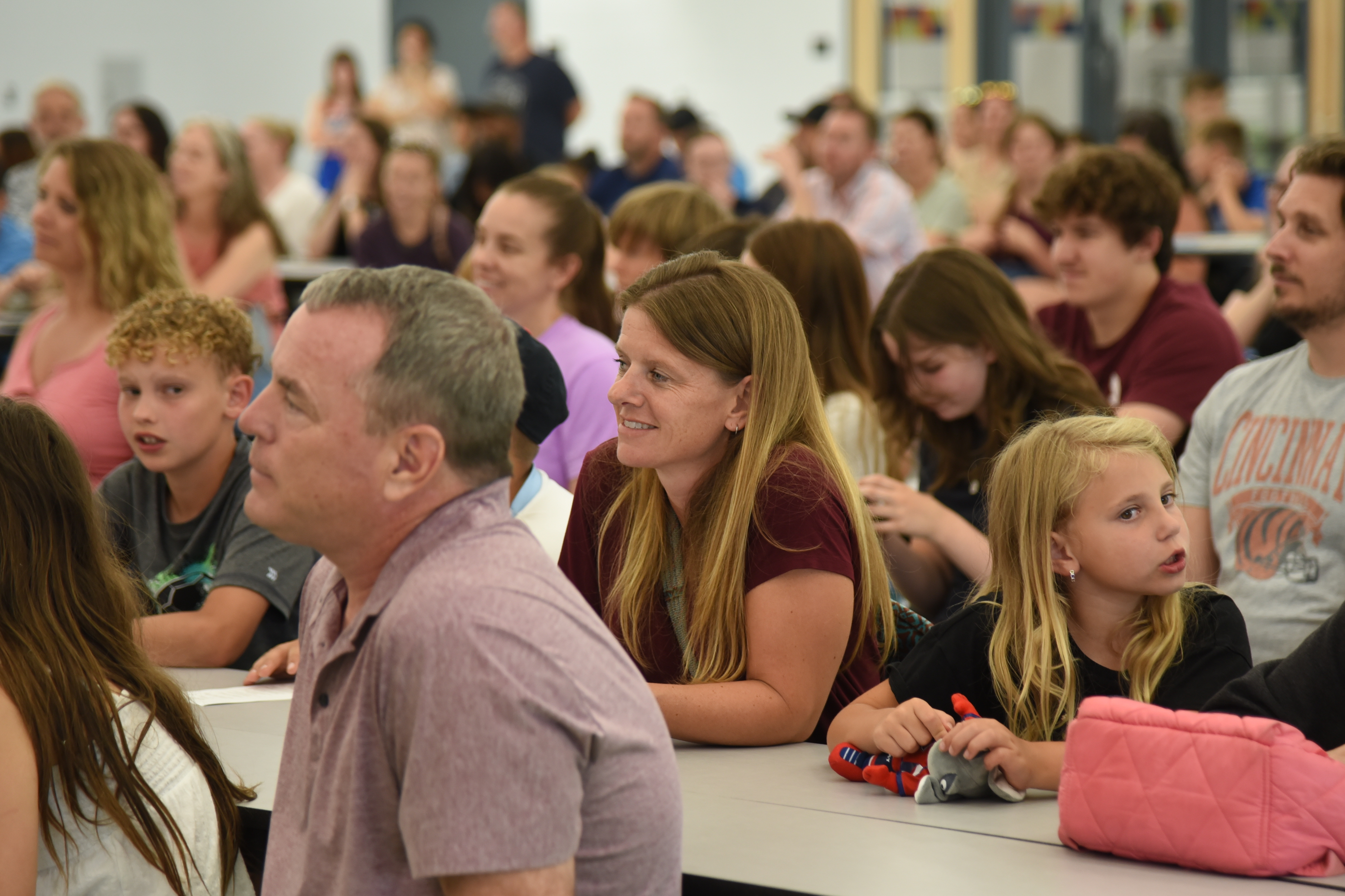 Large group of adults and children sitting at desks, facing front. Many are looking up, listening.