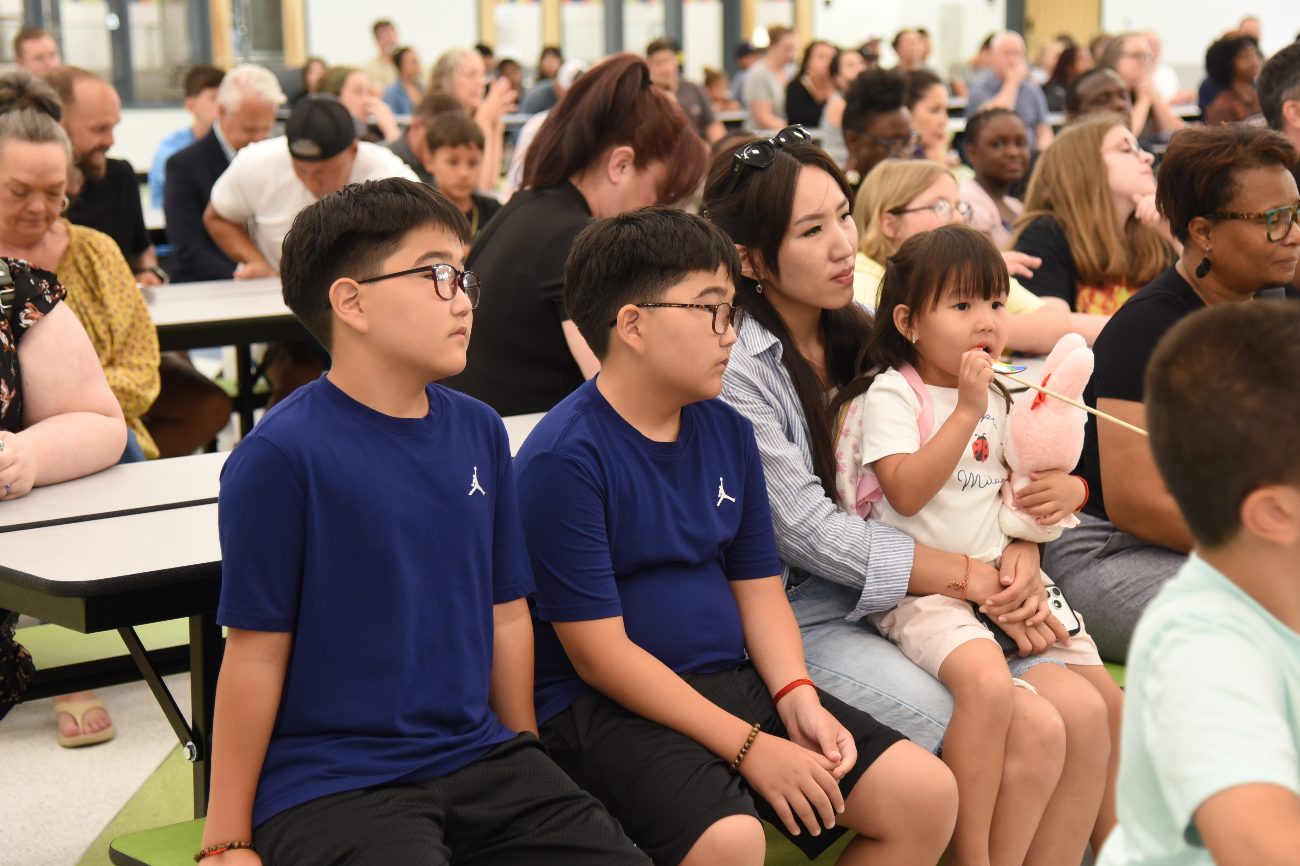 Group of people seated at tables. Some children and adults wearing glasses. Background: People seated at tables.