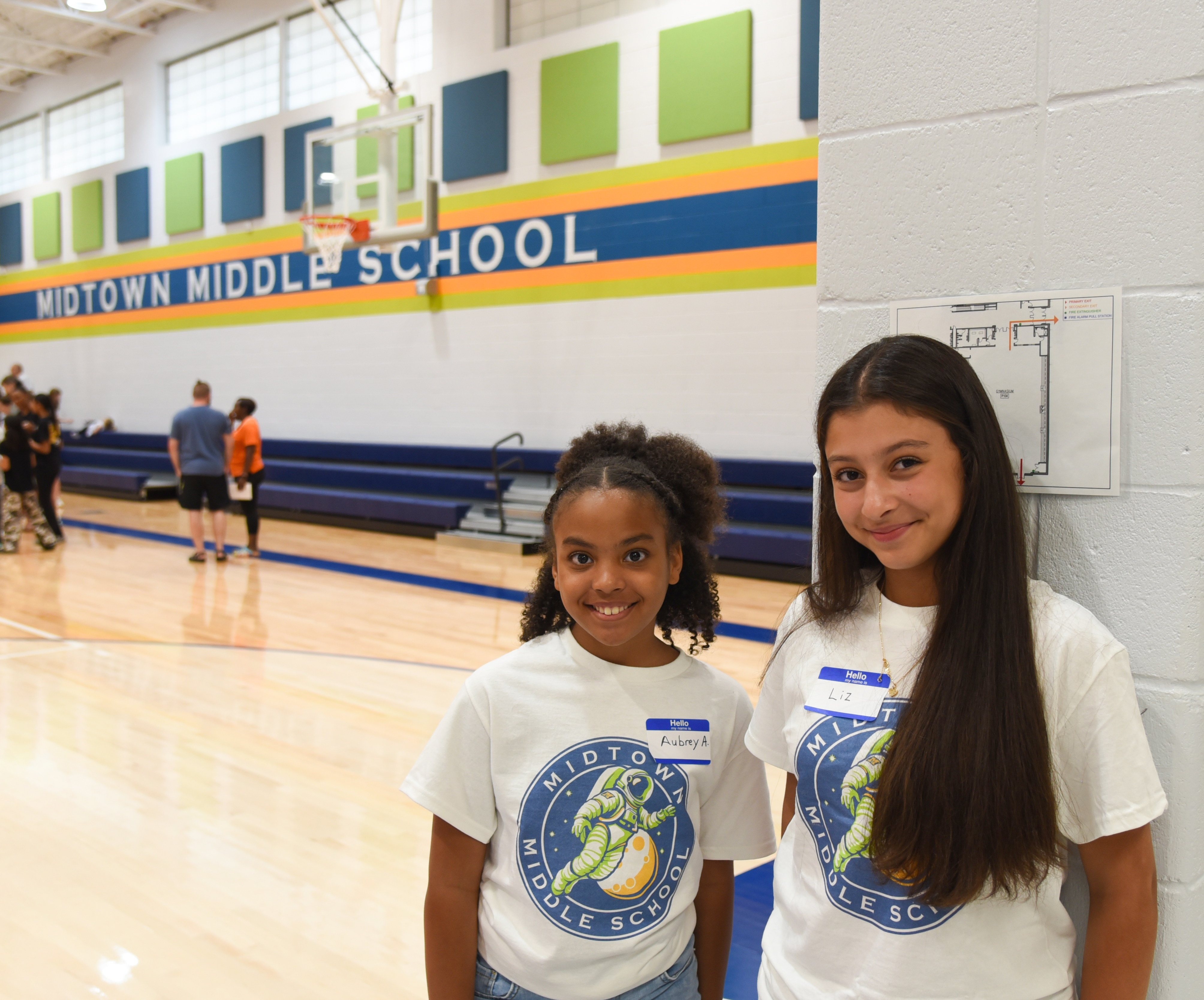 Two girls stand in a gym, smiling, with a sign reading "MIDTOWN MIDDLE SCHOOL" behind them.