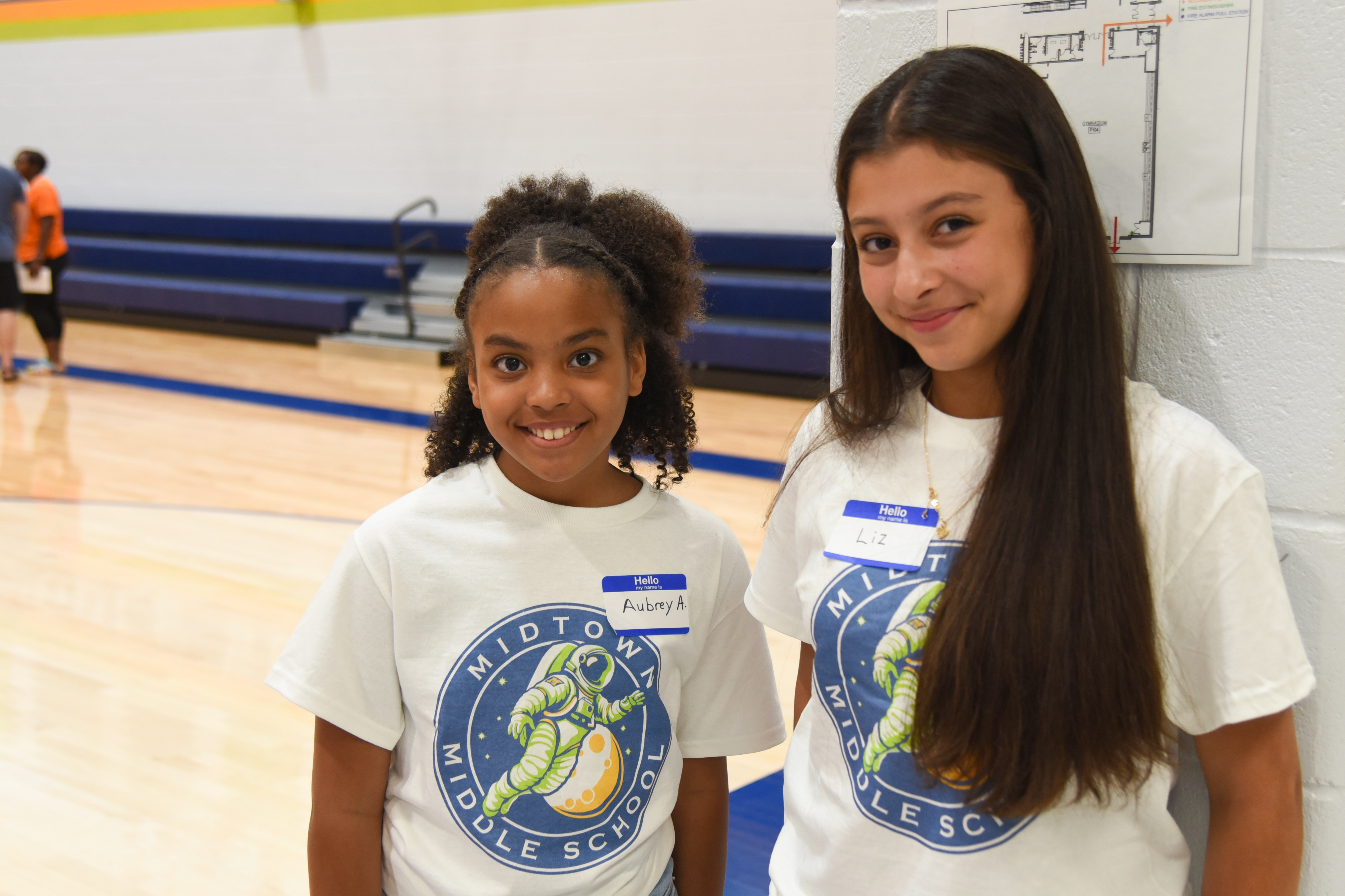 Two girls wearing matching white t-shirts with school logos stand together in a school gym. Behind them, a blue bench and a basketball court are visible.