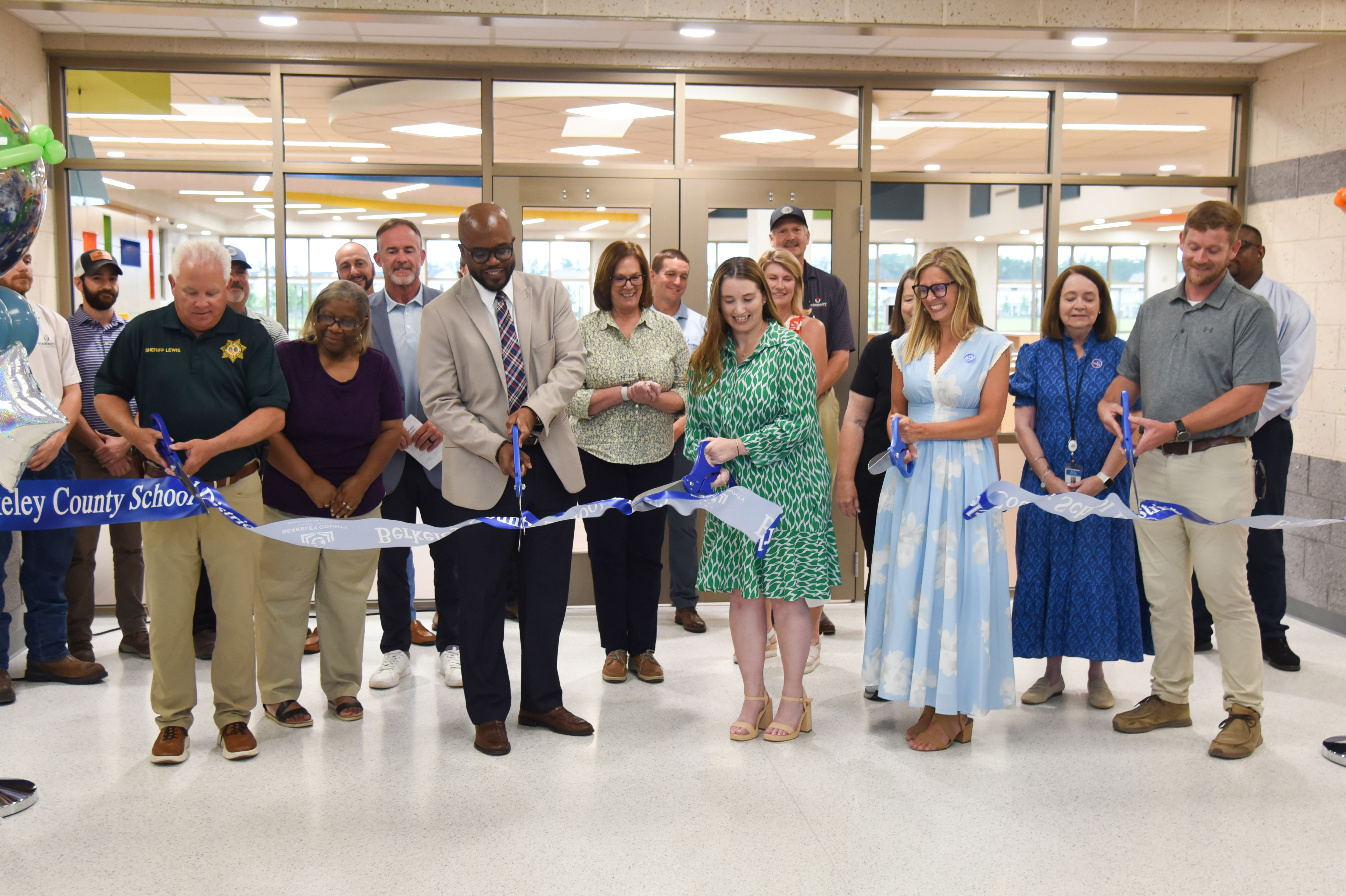Group of adults standing around a ribbon, with two holding scissors. One person wears a green dress.