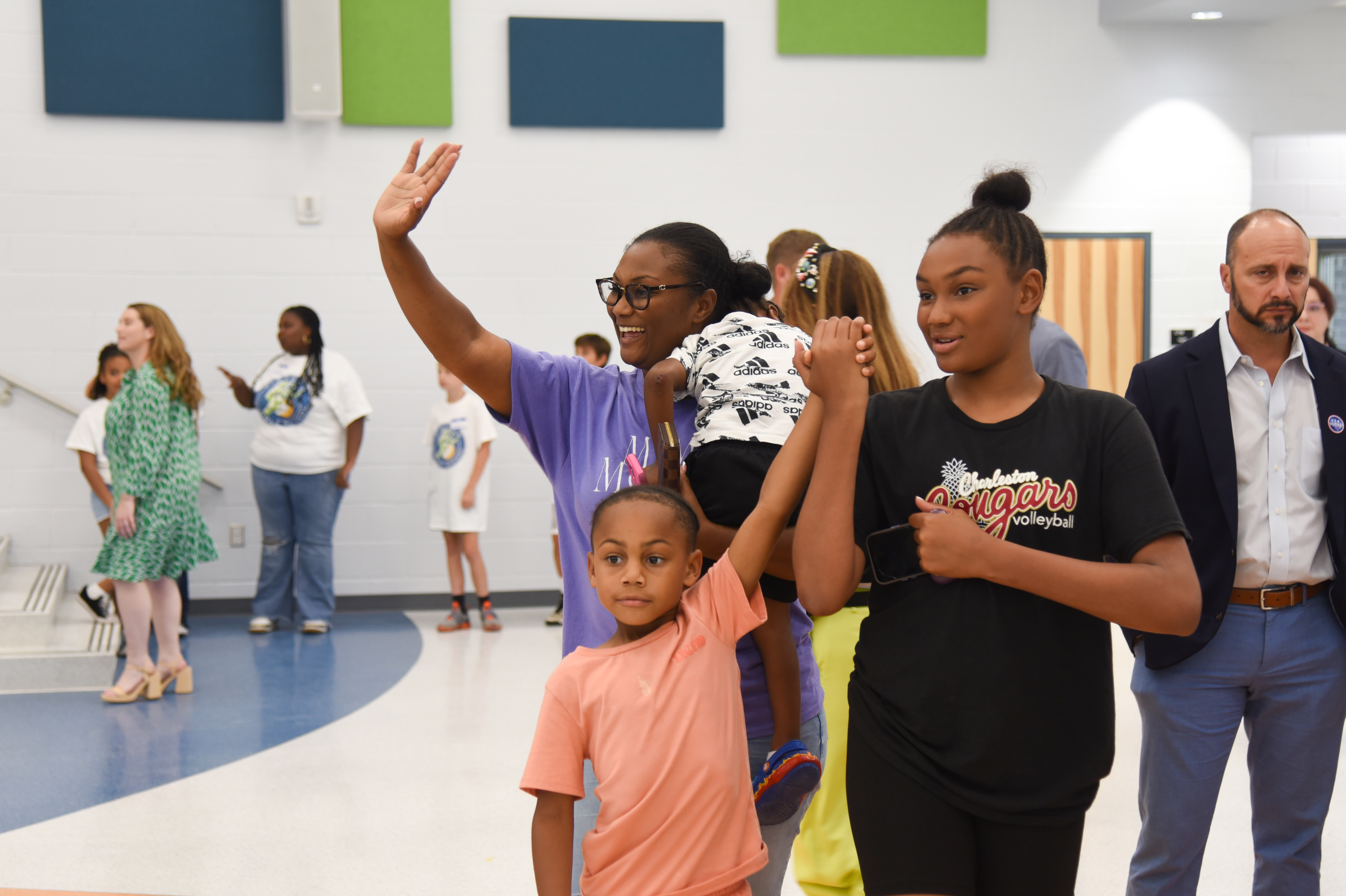 A group of people in an indoor gymnasium. A woman holds a child's hand, and another holds a baby.
