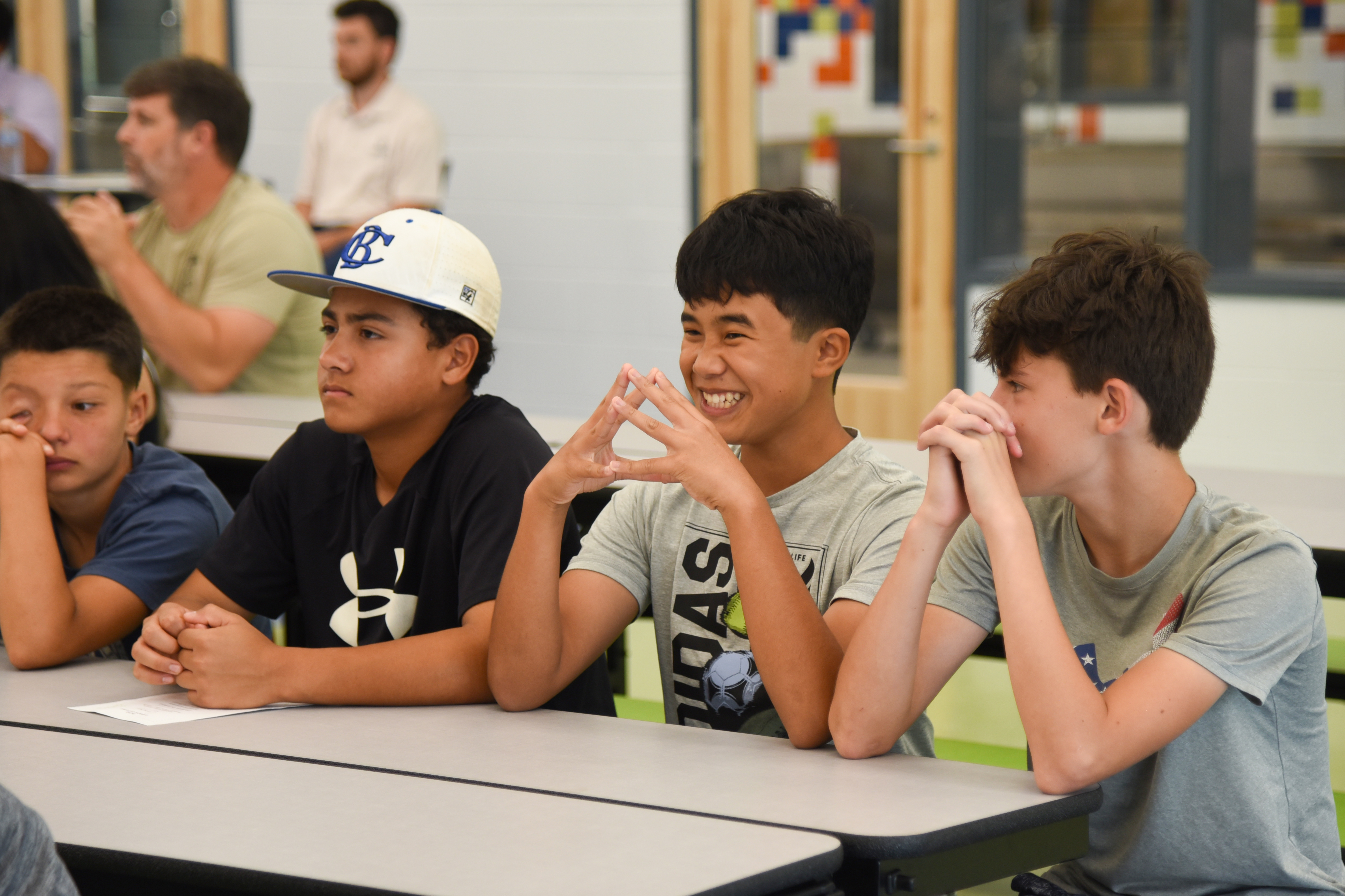 Four boys seated at a table, two smiling at each other, one making a heart sign, and one with his hand over his mouth.