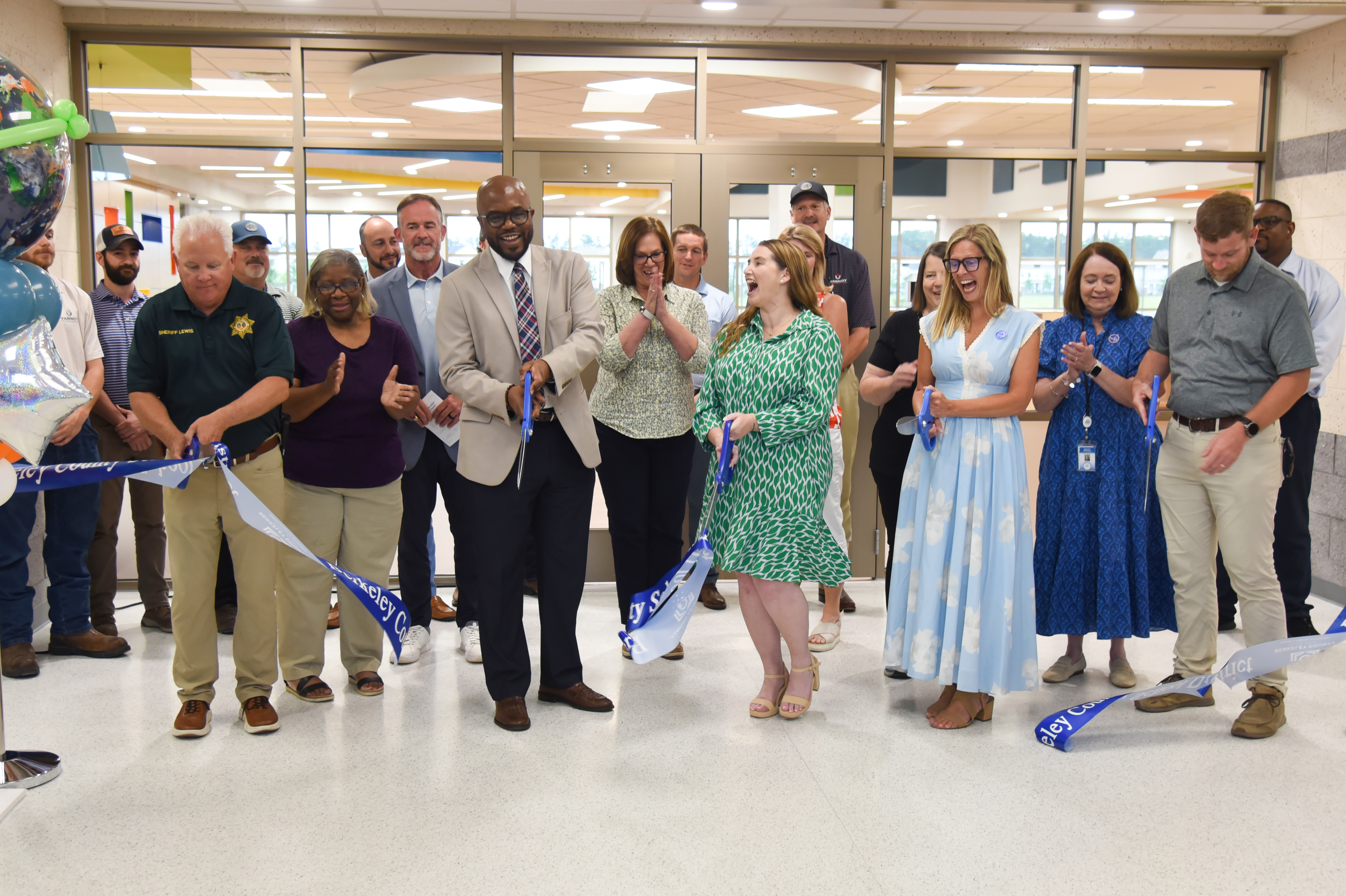 A group of people standing in a hallway, cutting a blue ribbon. They are wearing business attire.
