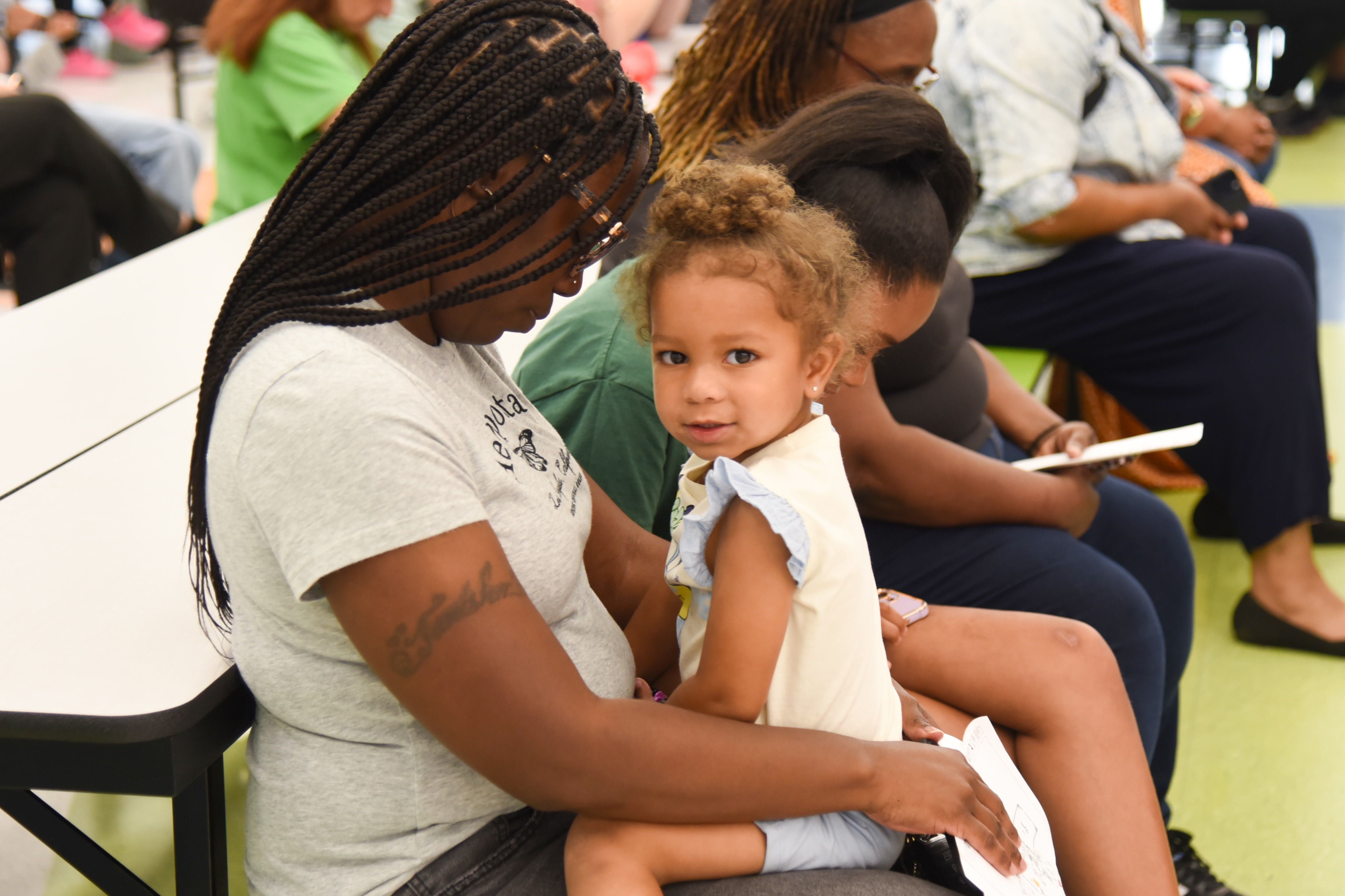 Woman holding a child, seated with others on benches in a room with white walls and green flooring.