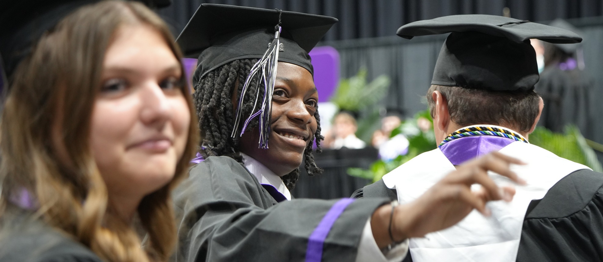 A woman in a graduation gown smiles at another woman while a man in a robe stands behind her.