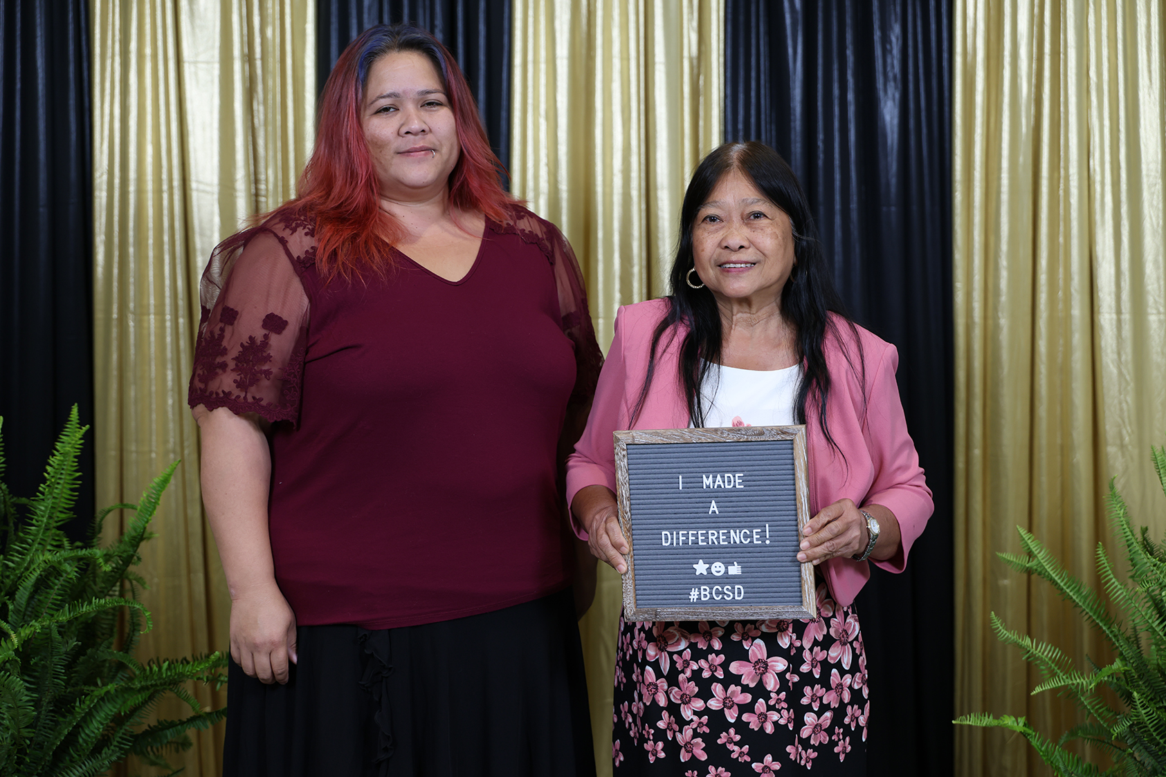 Two women stand with a sign that reads "I made a difference". The woman on the left wears maroon; the other wears a pink blazer.