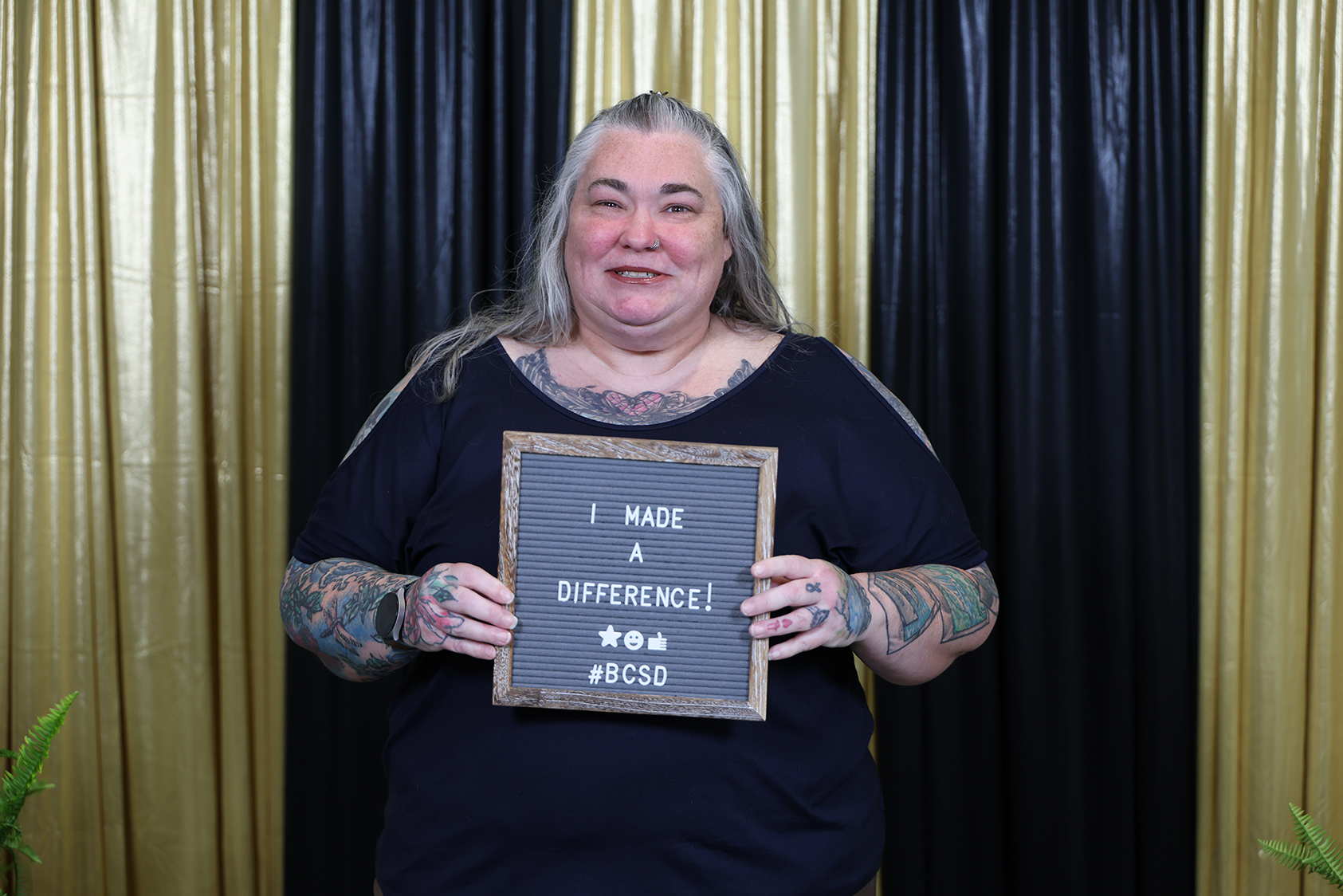 A woman holds a sign saying "I MADE A DIFFERENCE" in front of a black and gold curtain.
