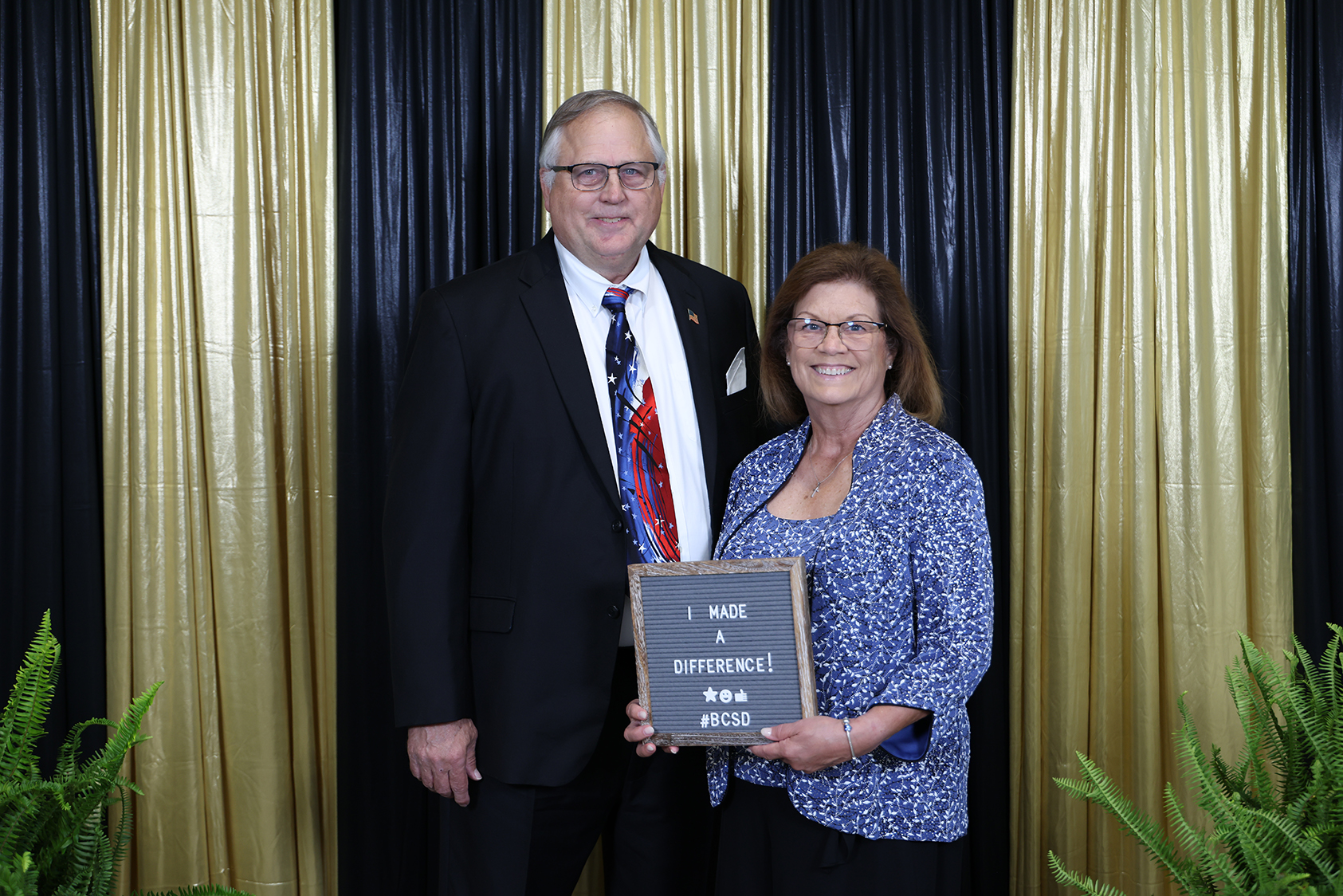 Two people stand in front of a curtained backdrop. The woman holds a plaque.
