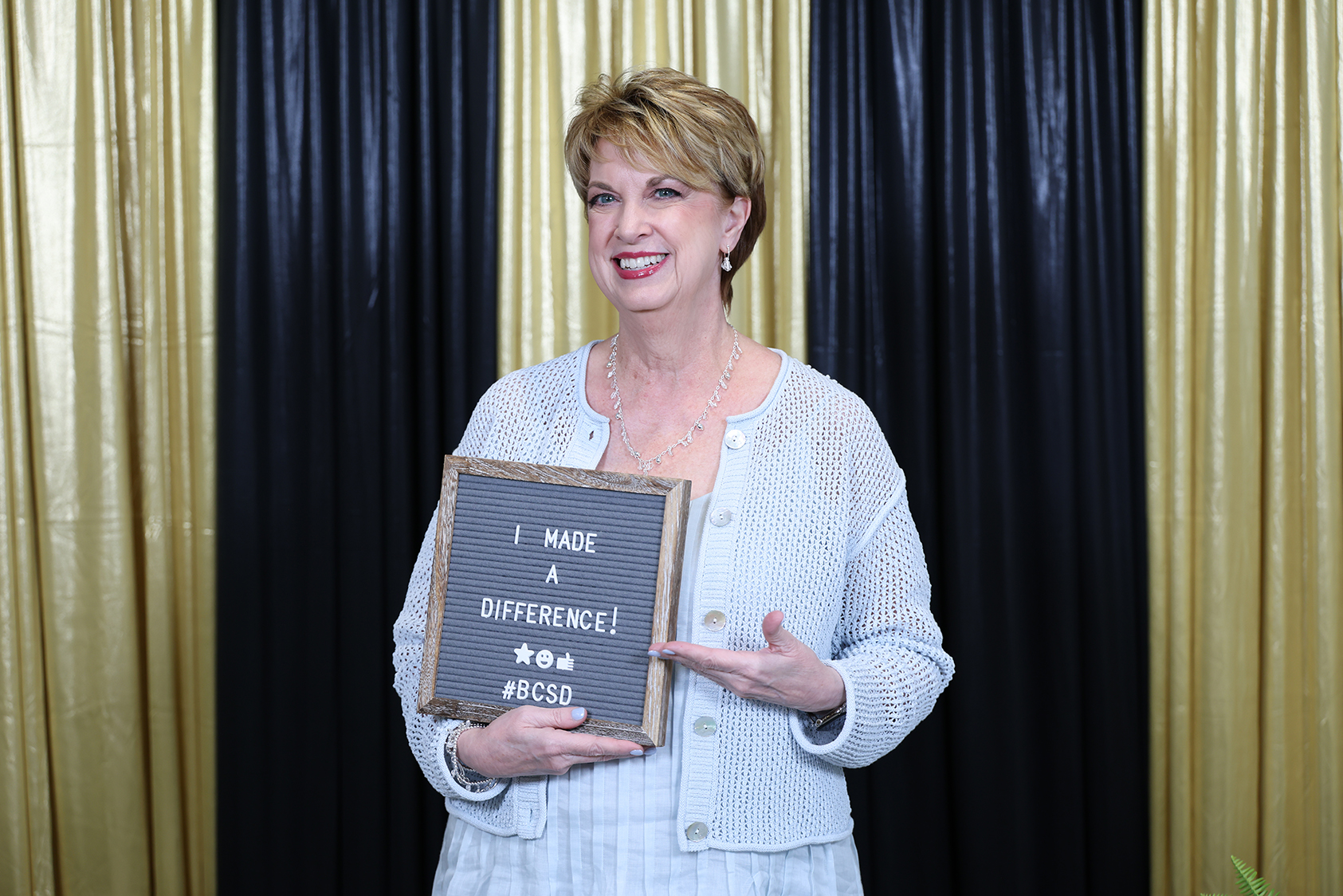 A woman with short hair smiles, holding a sign that reads "I made a difference!" against a gold-black curtain backdrop.