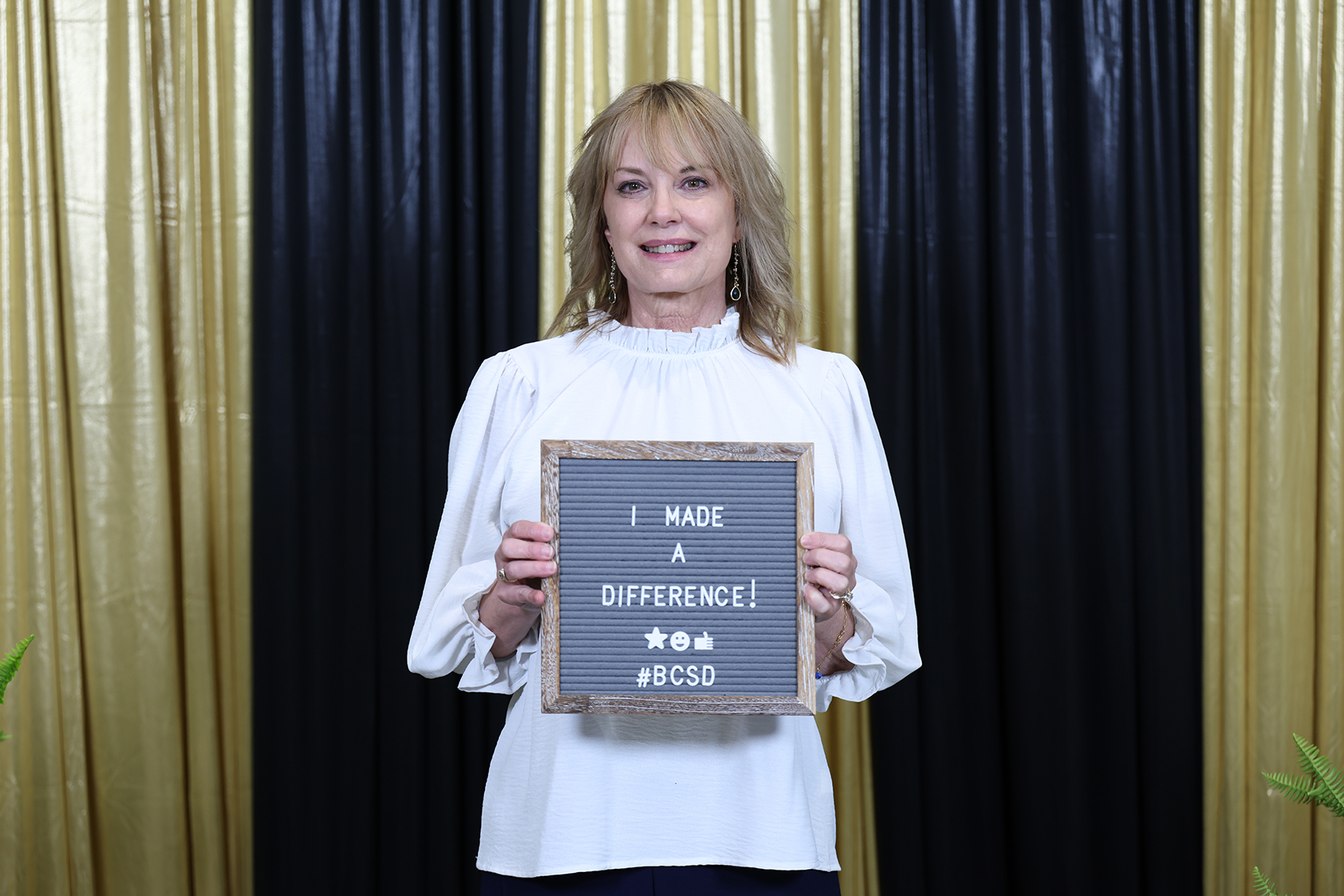 Person with blonde hair, holding a sign reading "I MADE A DIFFERENCE!" in front of a black and gold backdrop.