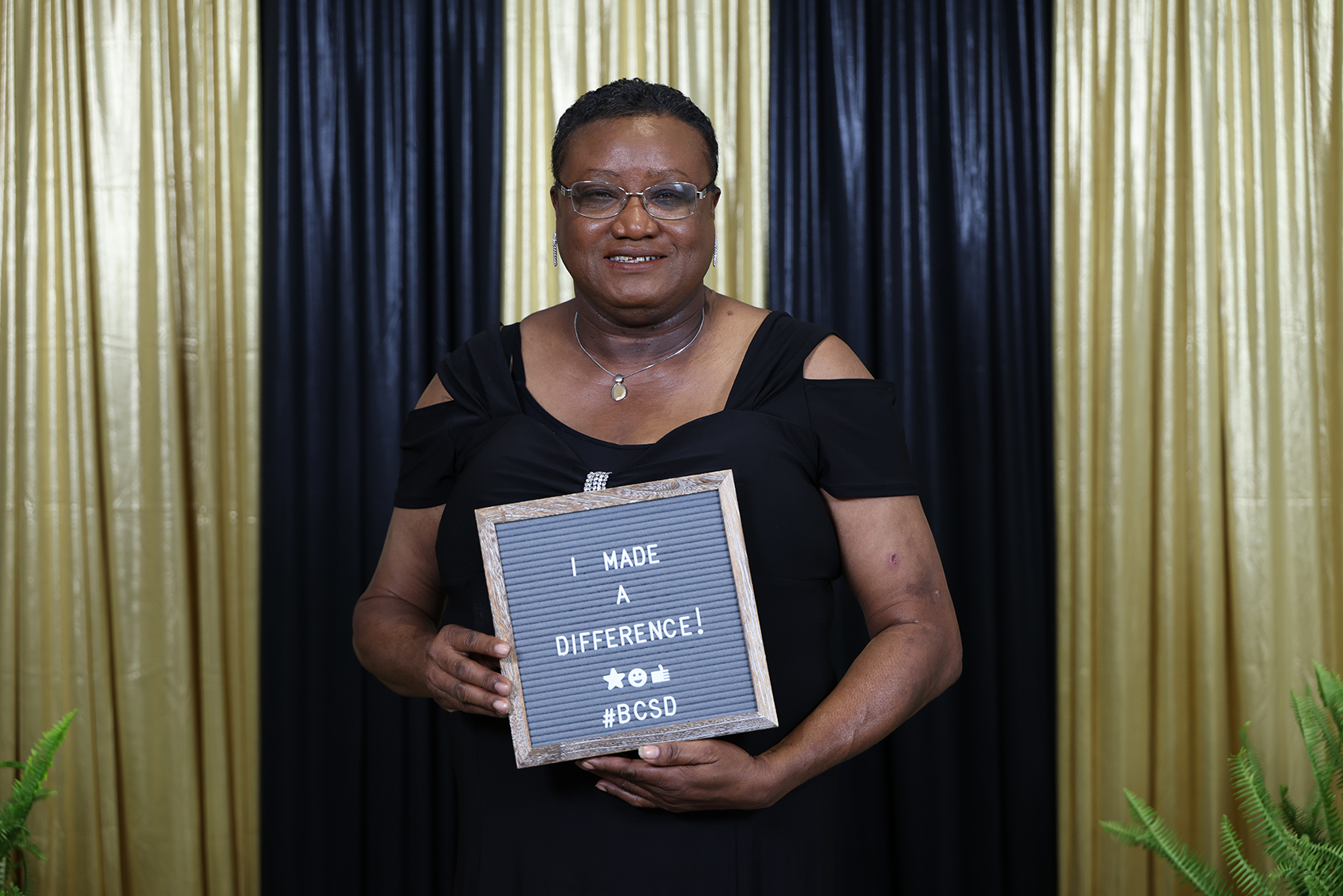 Woman in black dress, glasses, and necklace holds sign with "I Made A Difference!" text against a dark and gold curtain backdrop.