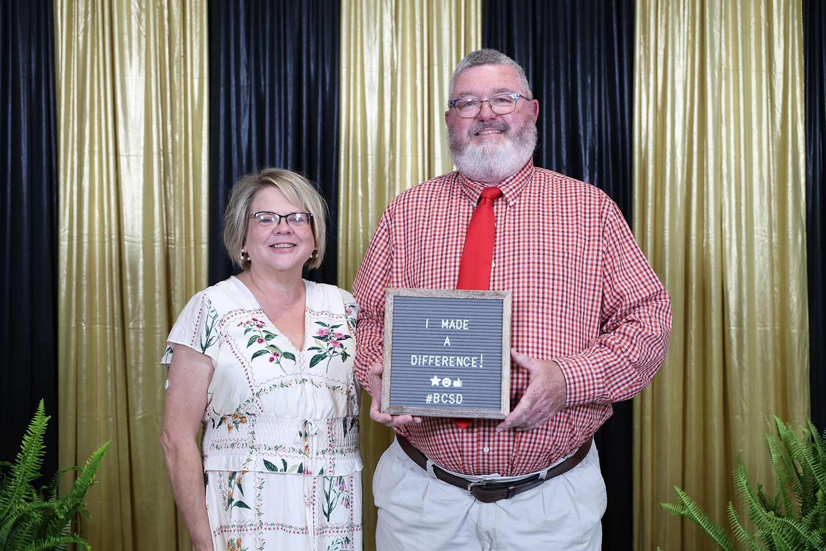 Man and woman stand behind golden curtains, smiling. Man holds plaque, woman wears floral dress.