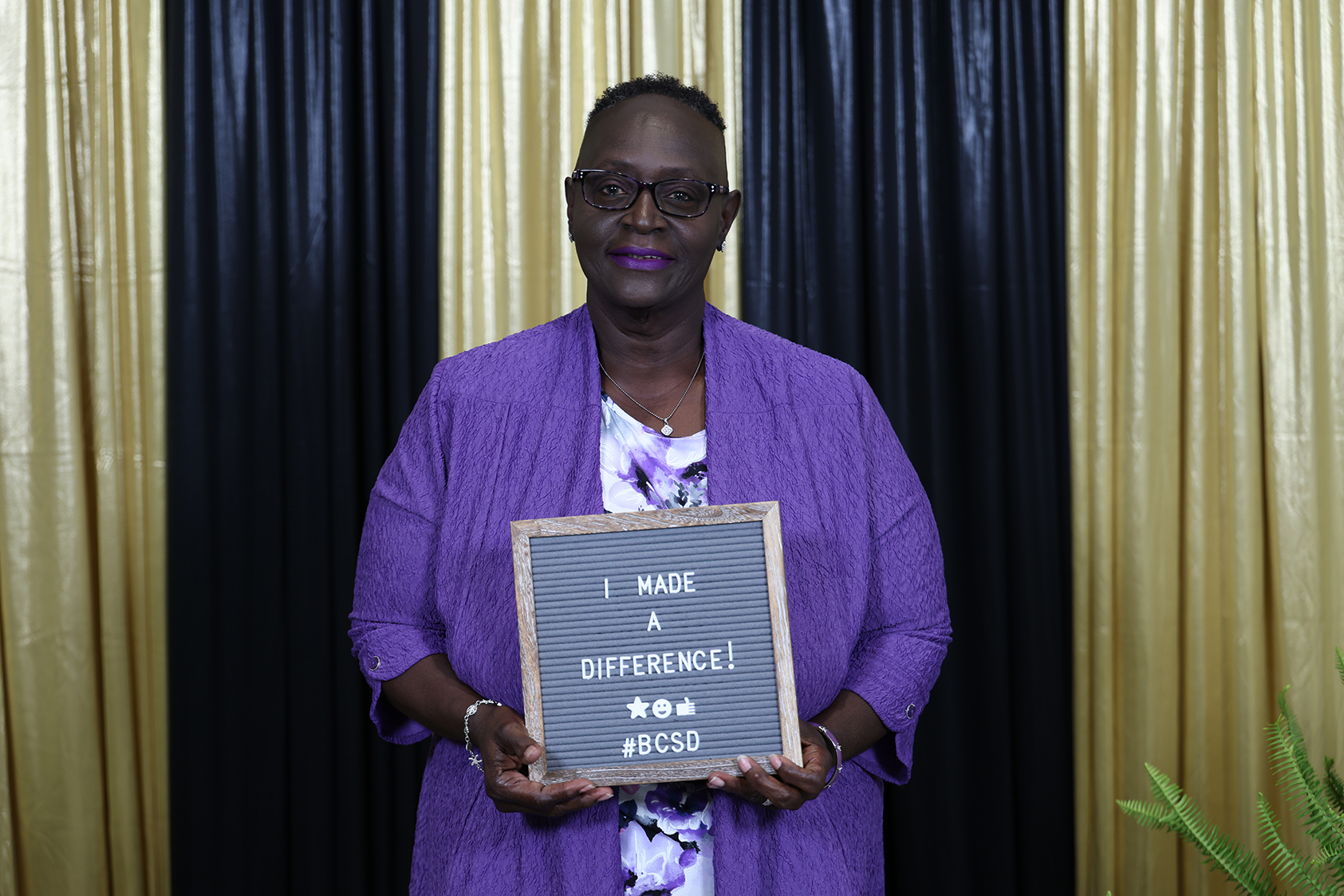 Person in purple holds a chalkboard sign with "I MADE A DIFFERENCE" in front of black and gold curtains.