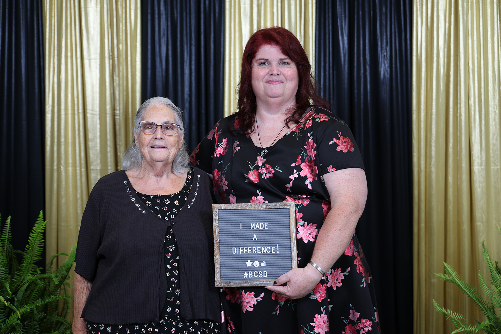 Two women pose with a chalkboard plaque reading "I Made a Difference" against a black and gold backdrop.
