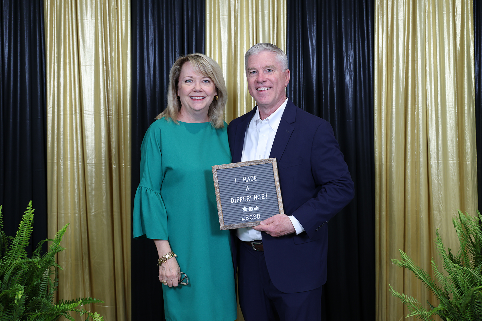 A man and a woman stand together, smiling, in front of a striped backdrop, holding an award.