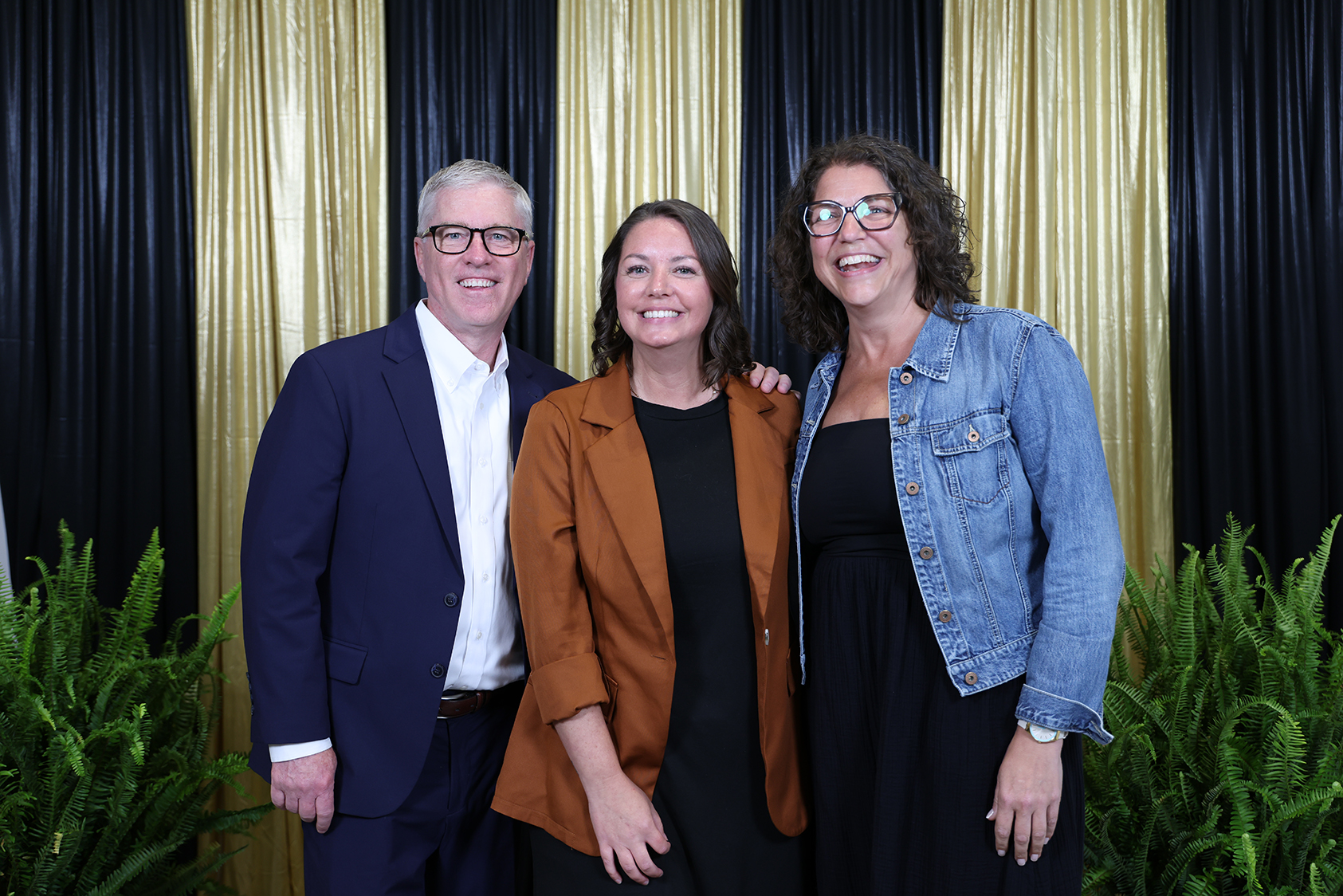 Three people standing together in front of a black and gold backdrop with plants.
