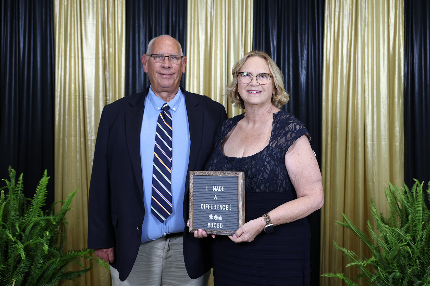 A man and a woman in formal attire stand together, smiling, with the woman holding a plaque.