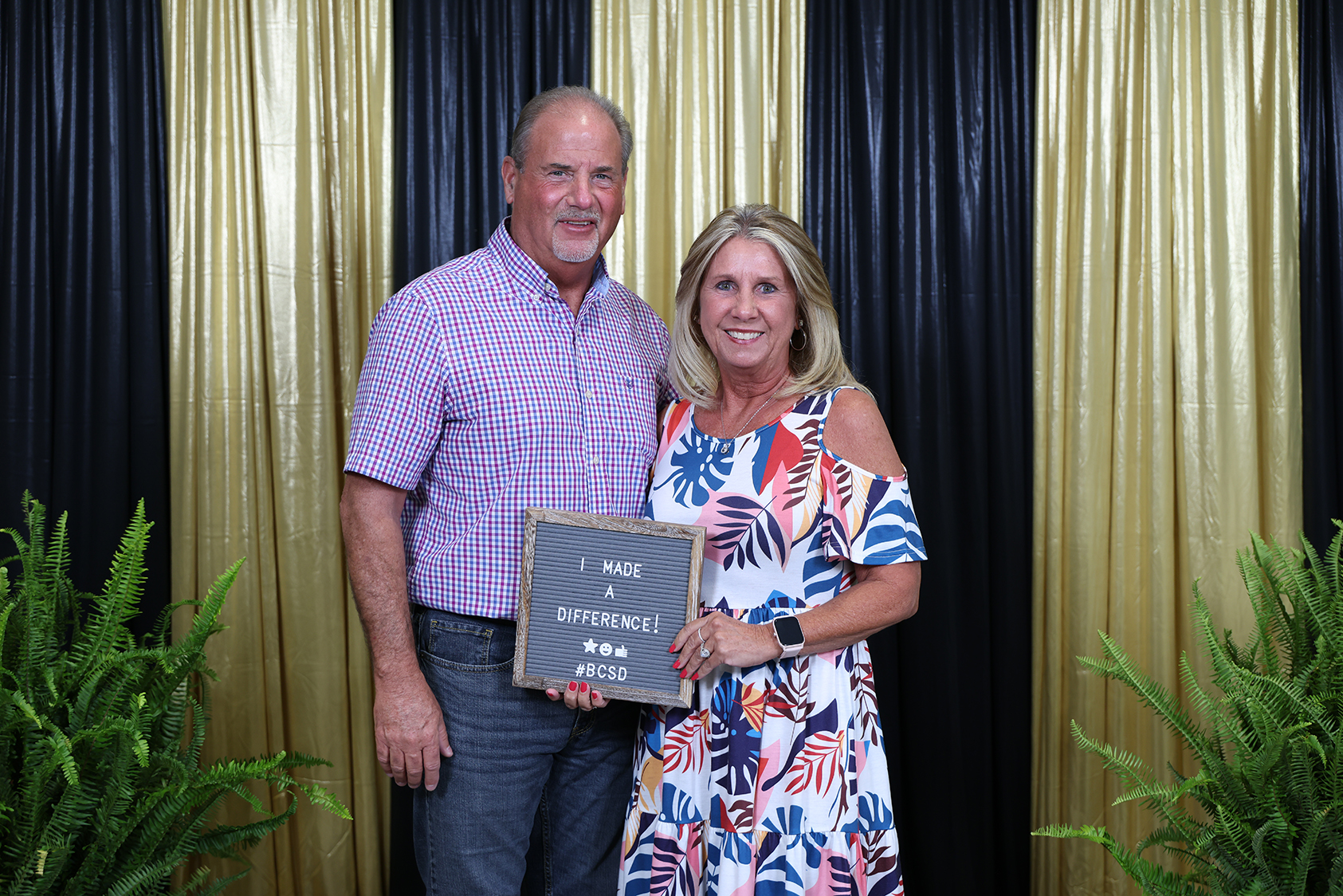 A man and a woman stand beside each other, holding a plaque. The man wears a pink shirt, and the woman a colorful dress.