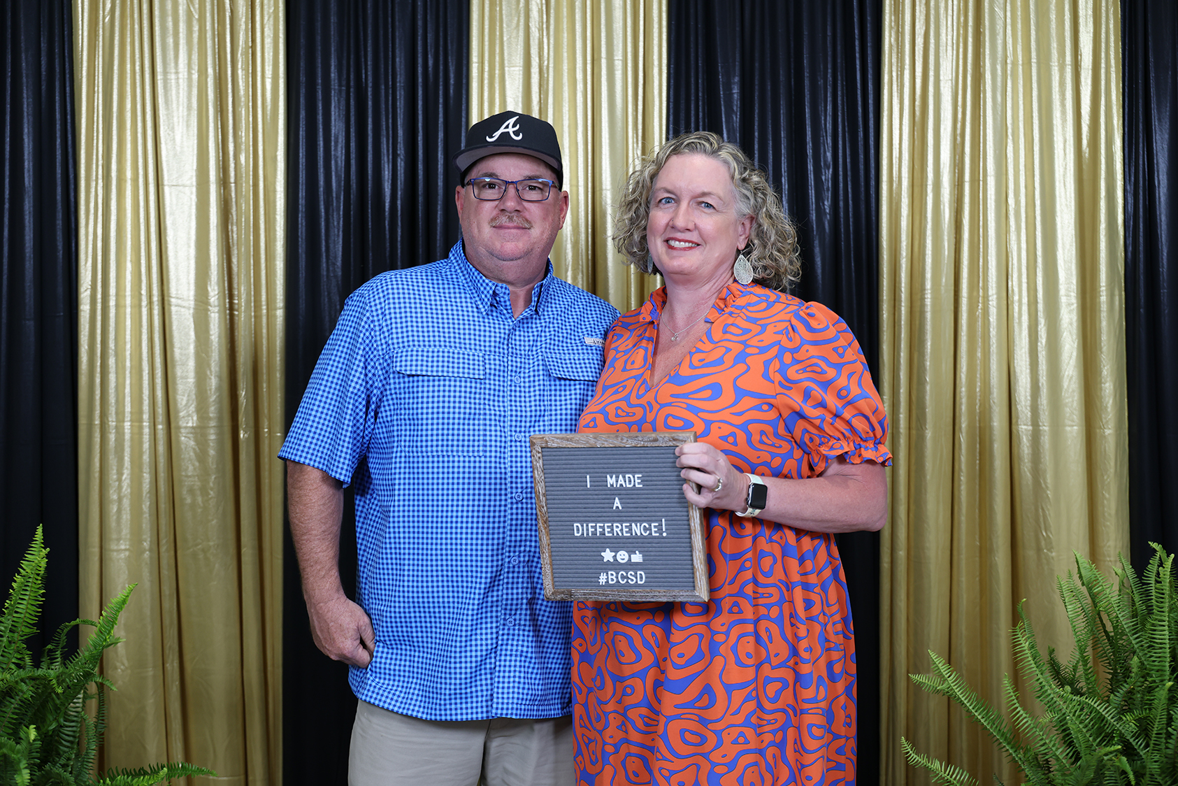 Man and woman in orange and blue clothing holding a sign, standing before a black and gold striped curtain.
