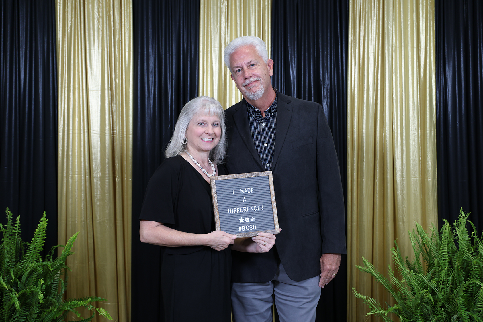 A man and a woman stand together in front of a black and gold backdrop. They hold a framed award.