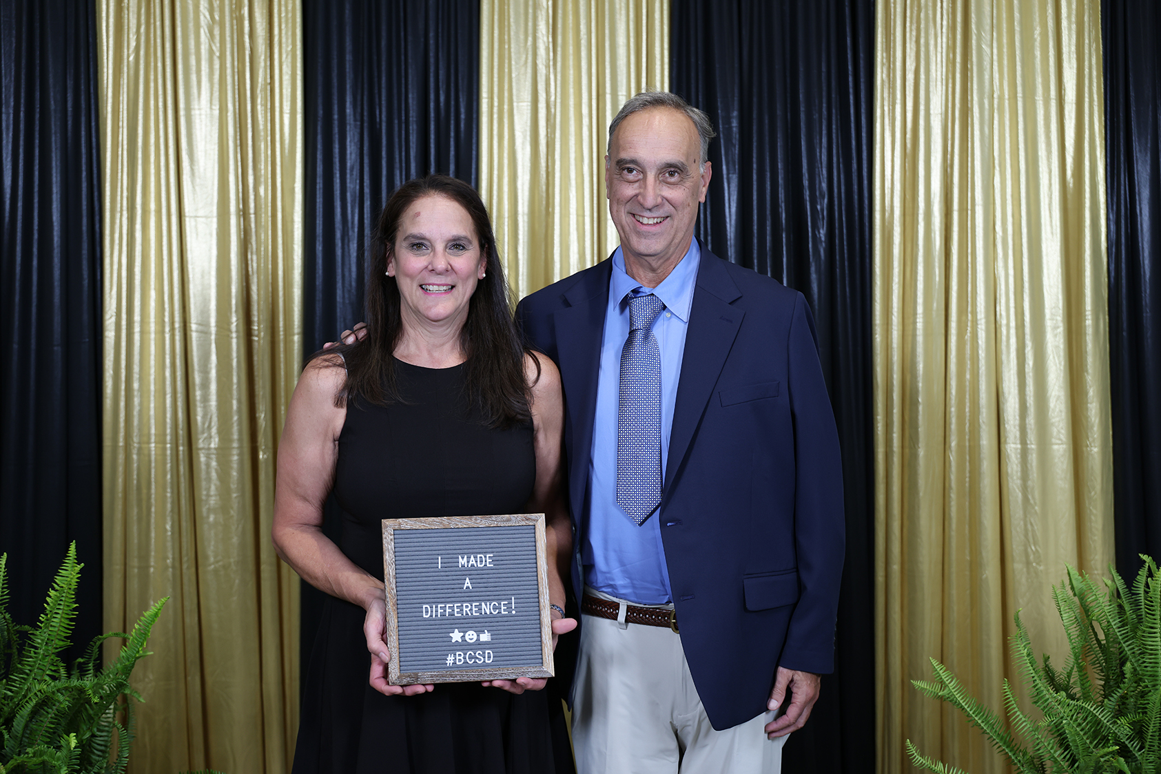 A woman holds a plaque reading "Made a Difference" with a man in formal attire standing behind her. They stand against a black and gold curtain.