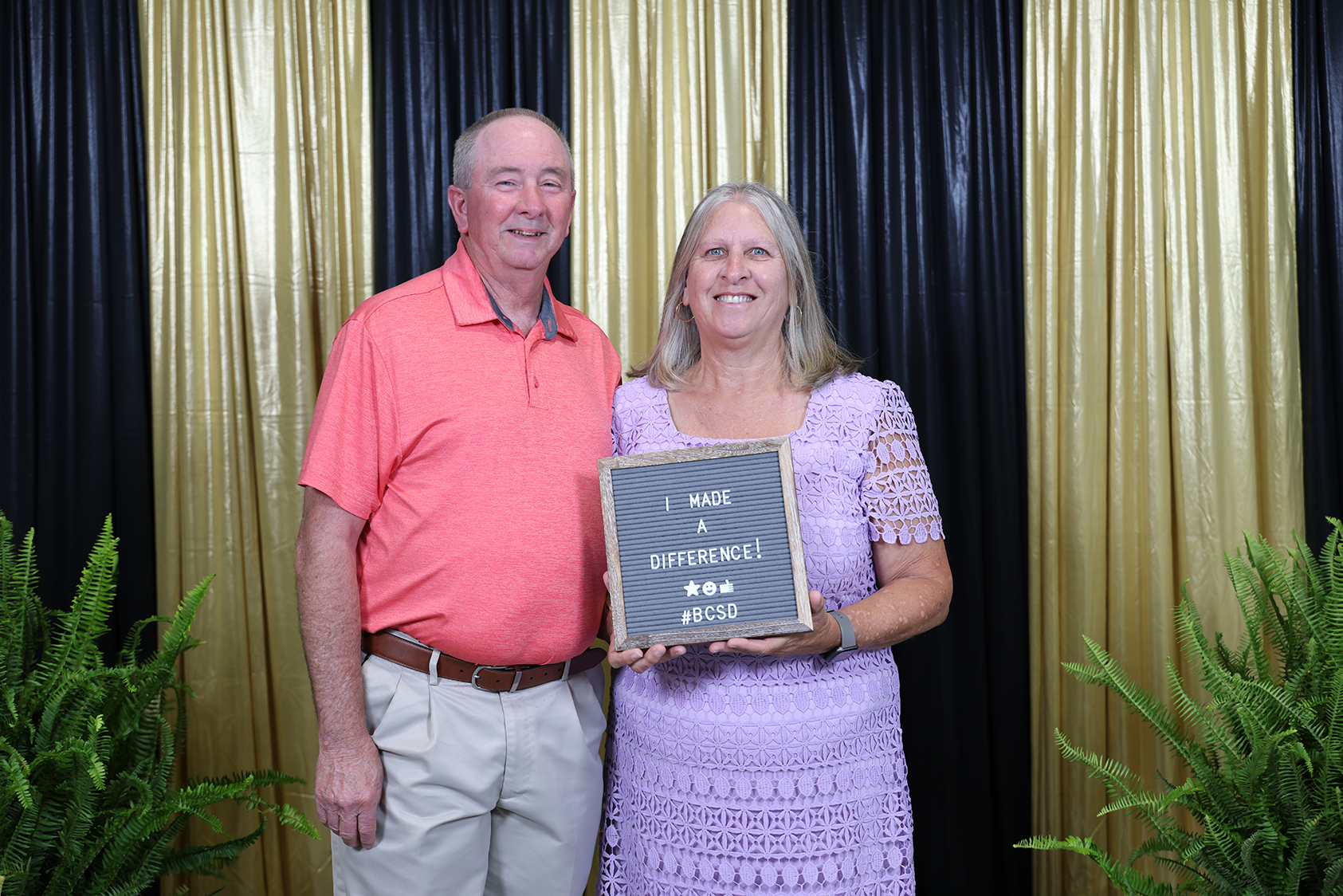 A man and woman stand with a plaque that says "I Made a Difference." They are in front of decorative curtains.