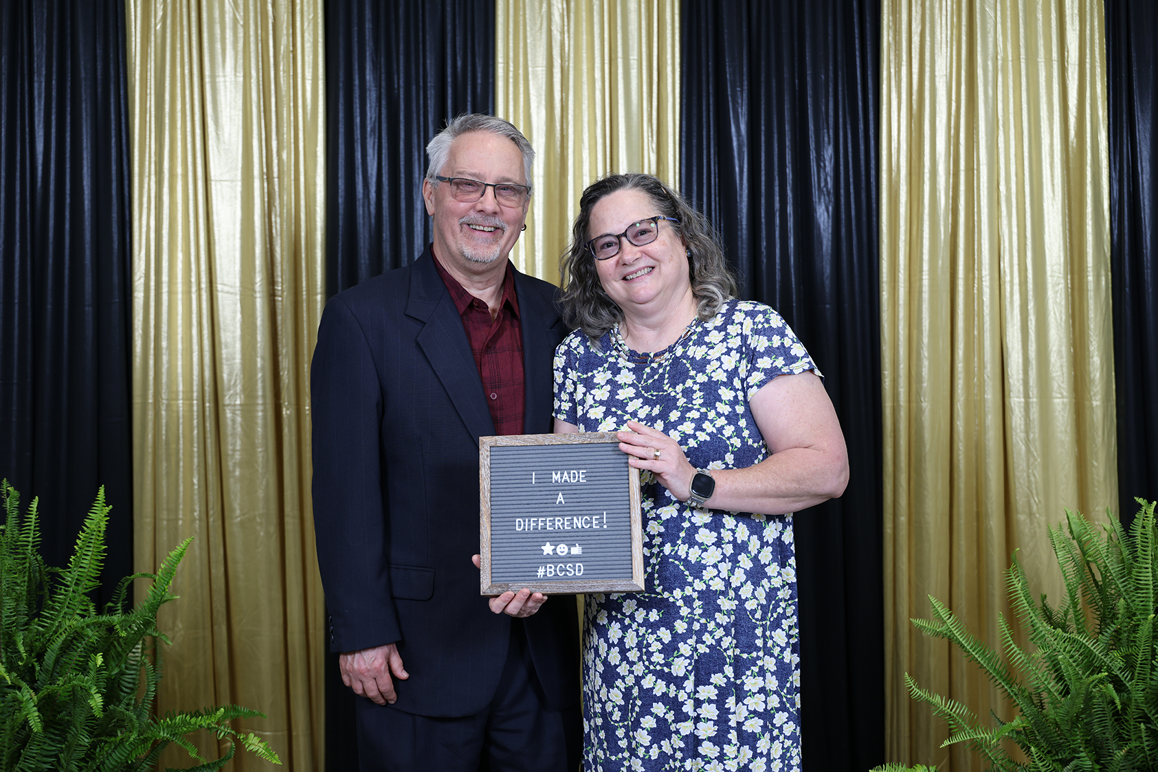 A man and a woman holding a framed plaque in front of a gold and black backdrop with plants.