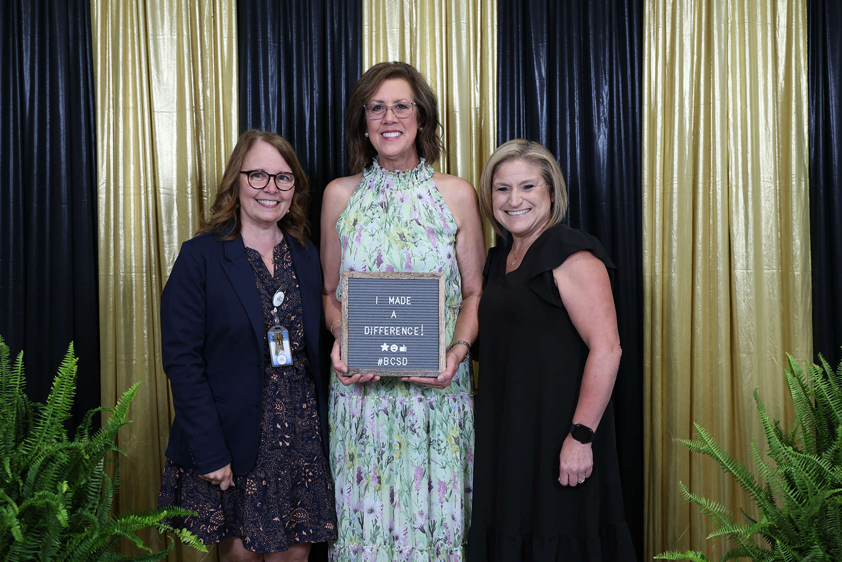 Three women stand with a woman holding a framed award. One wears a dress, another a blazer. They pose with a backdrop of gold and black curtains.