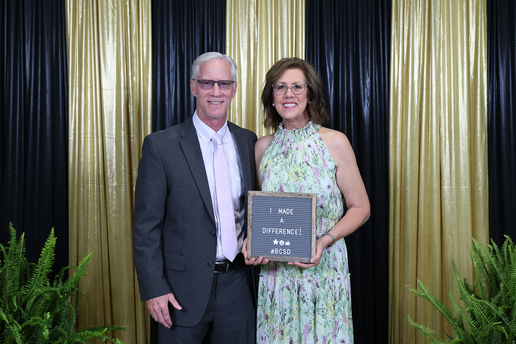 A man and woman stand together, holding a framed message about making a difference. They stand in front of gold curtains and plants.