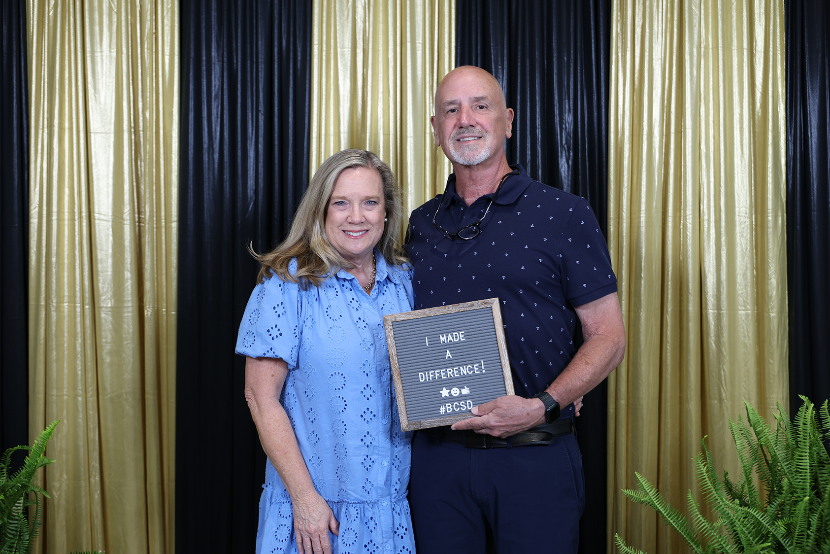 A man and woman stand in front of a black and gold backdrop holding a sign.