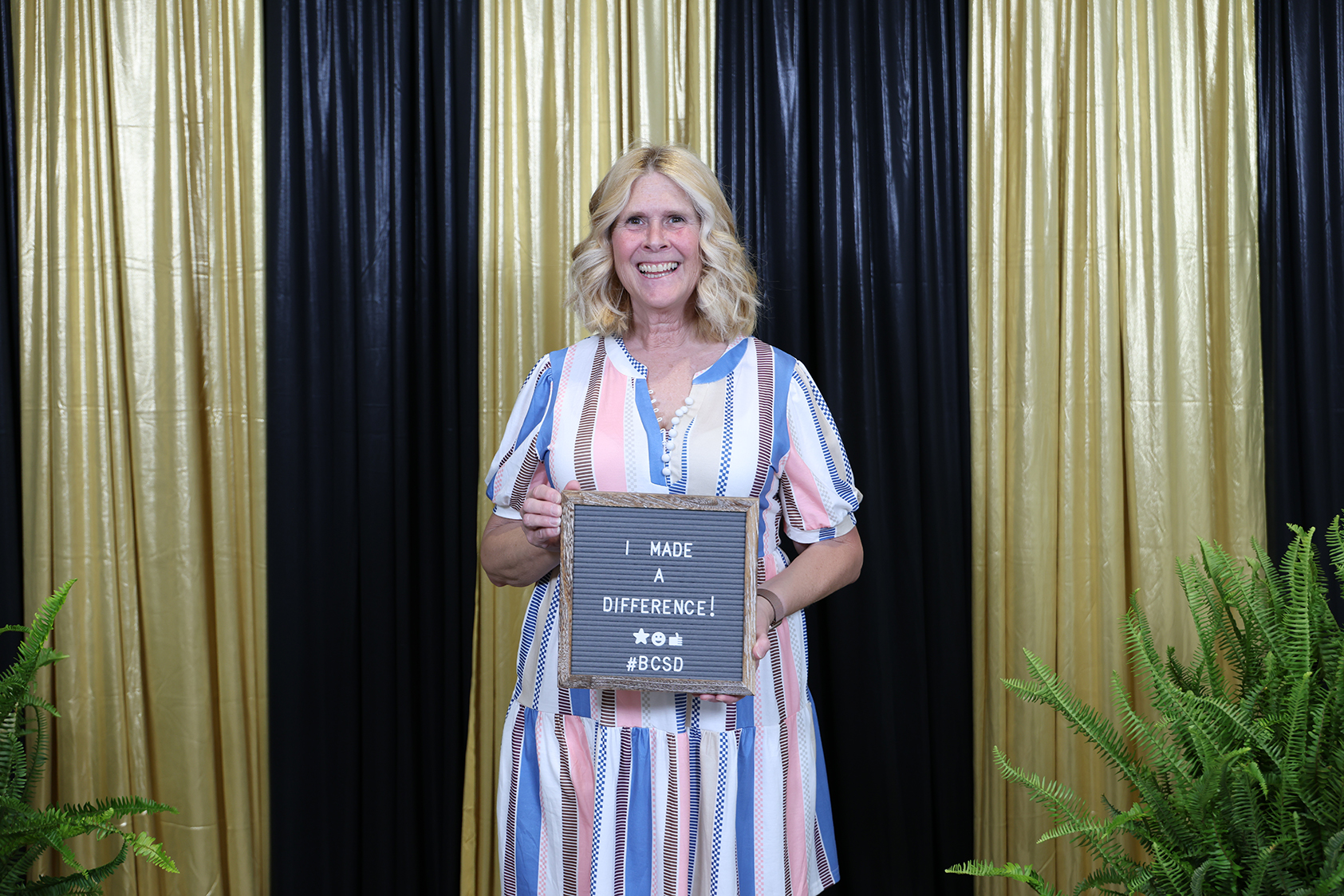 Woman in a multicolored dress holds a chalkboard that reads "I made a difference". Behind her are yellow and black curtains.