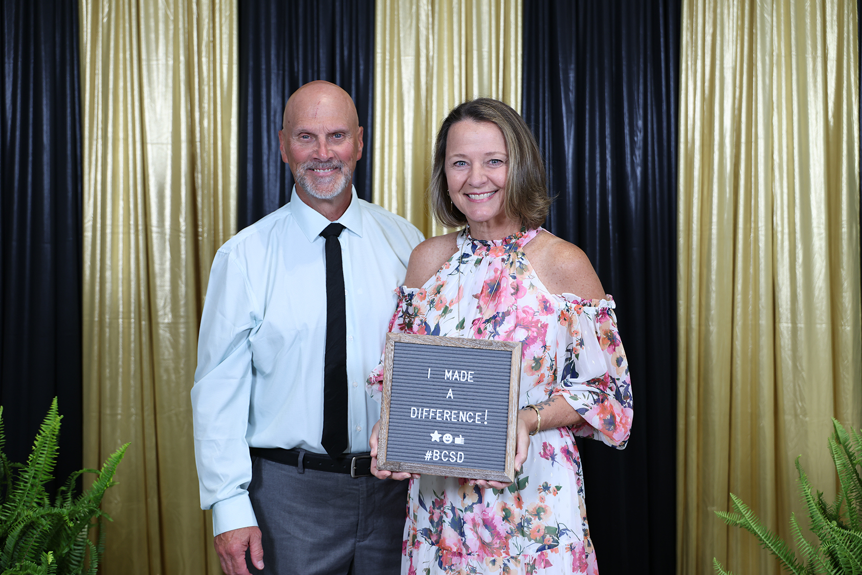 A man and woman stand together holding a plaque with the words "I made a difference." They are in front of a black and gold backdrop.