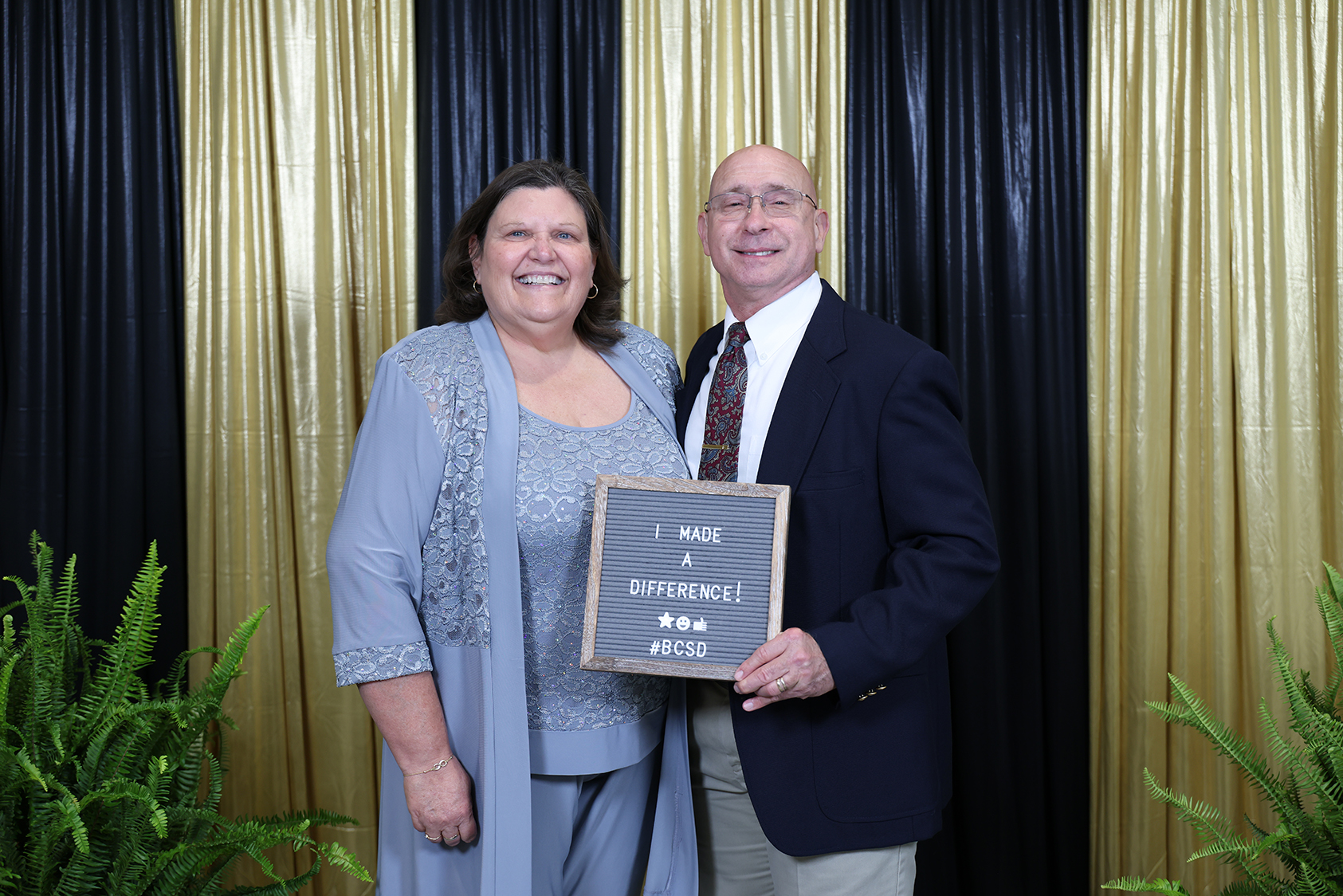 A man and a woman pose in front of a black and gold curtain, holding a plaque.