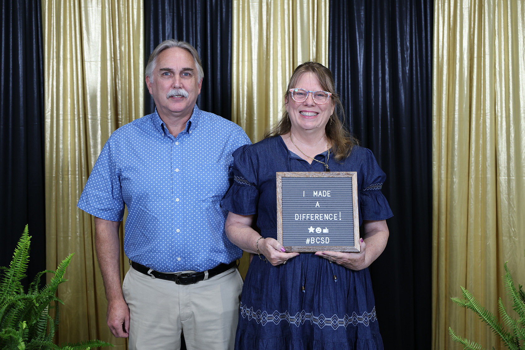 A man and a woman stand against a gold and black backdrop. She holds a plaque.