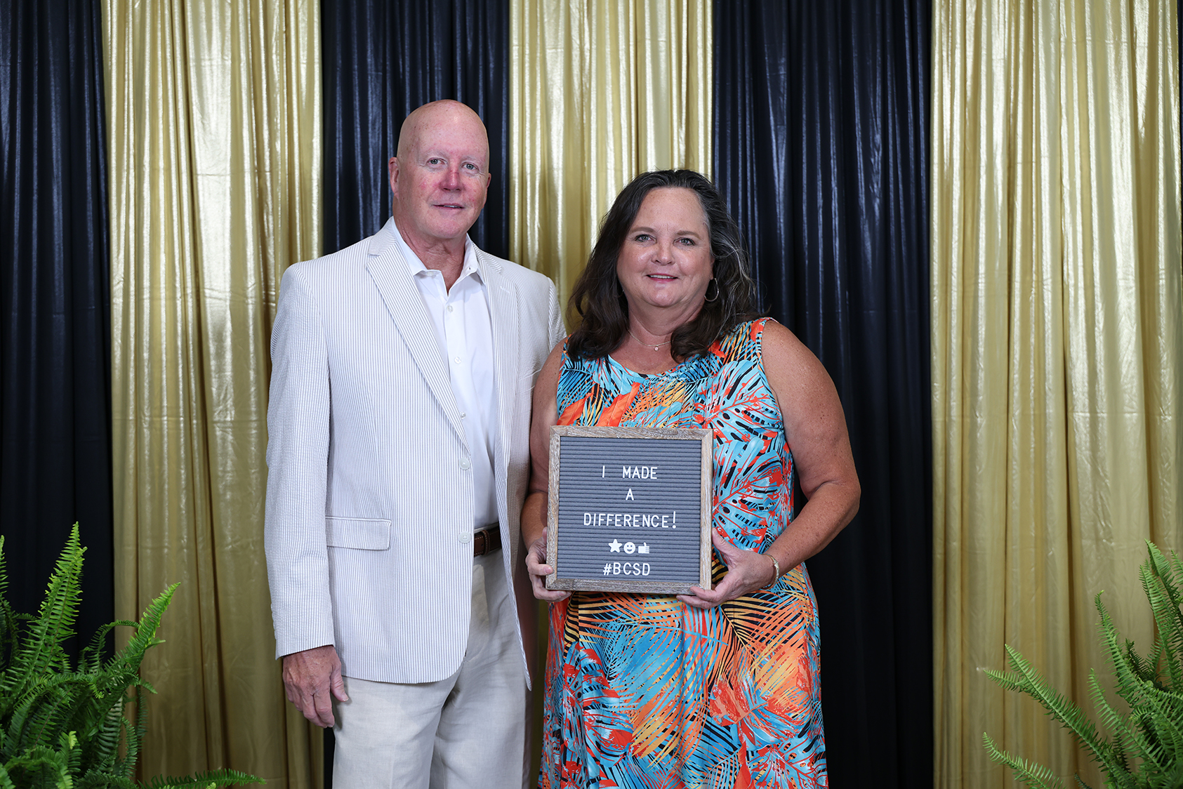 A man and a woman stand with a plaque that says "I Made a Difference." Behind them, black and gold curtains.