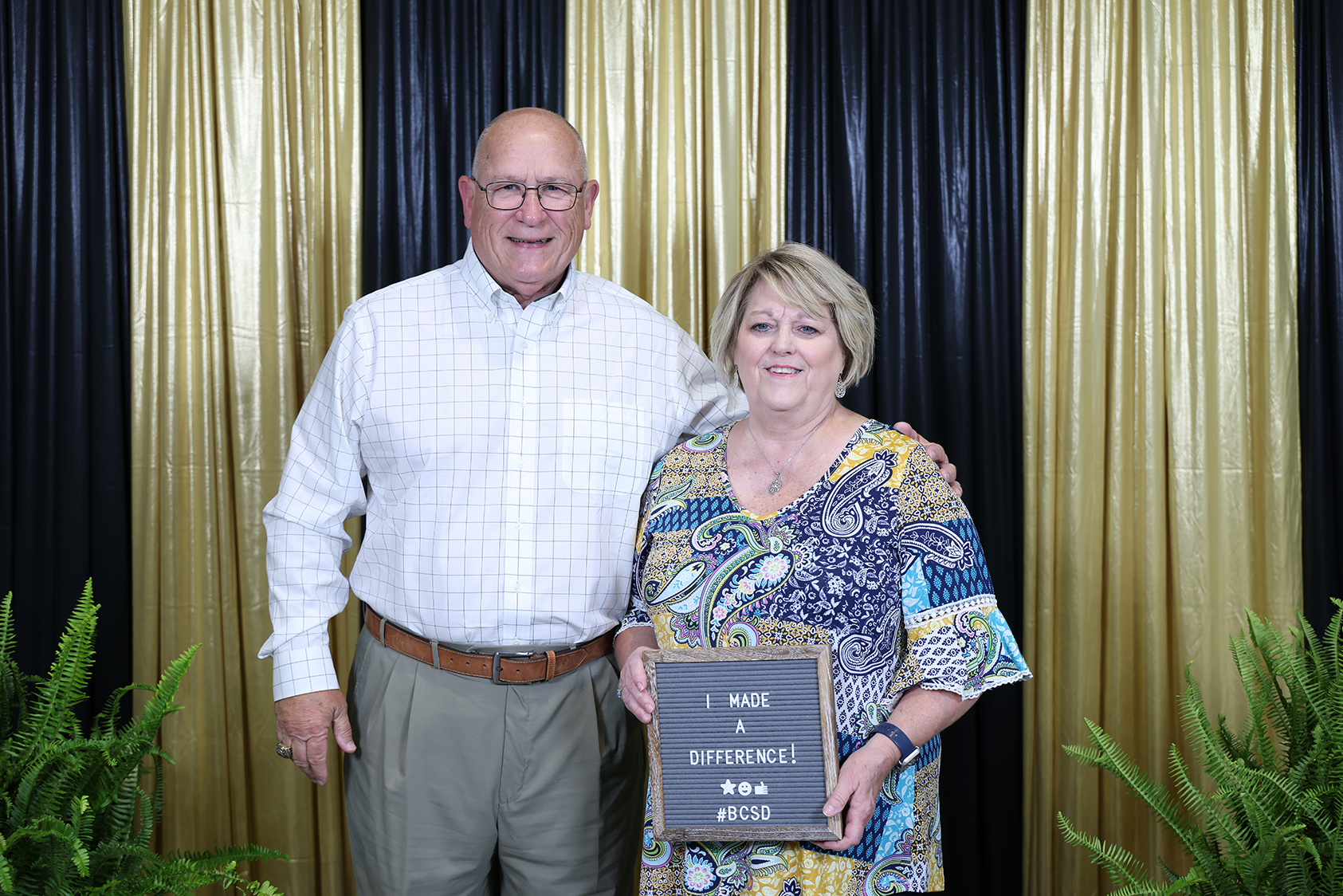 A man and a woman stand side by side, smiling. The woman holds a plaque with text. Behind them, a backdrop with golden curtains and greenery.