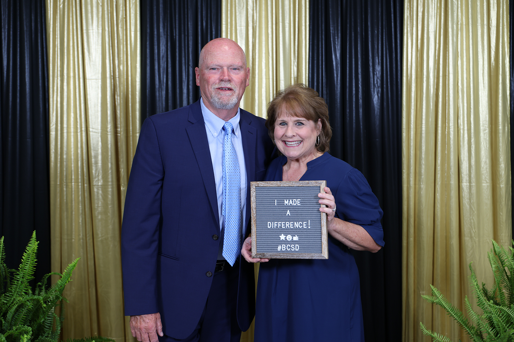Man and woman stand beside a photo backdrop. He wears a dark blue suit and tie. She wears a blue dress. They hold a plaque.