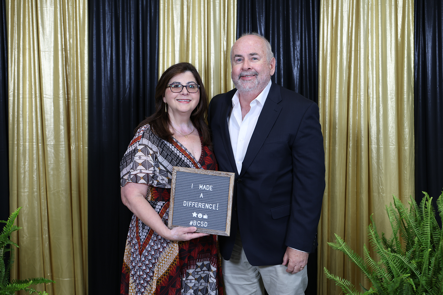 A man and a woman stand next to each other holding a plaque that reads "I Made a Difference." Behind them, gold and black curtains provide a backdrop.