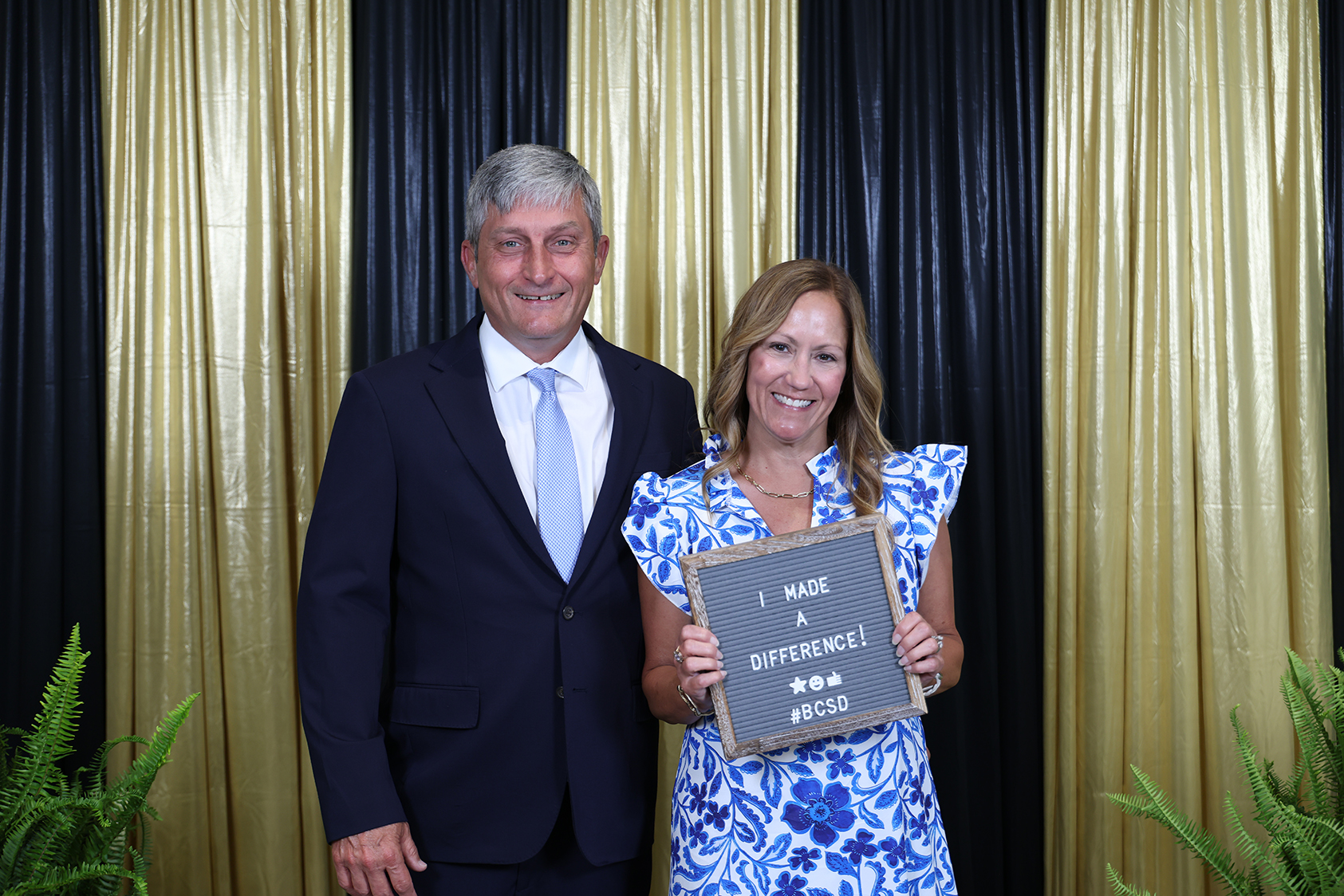 A man and woman stand behind a backdrop of black and gold curtains, holding a chalkboard.