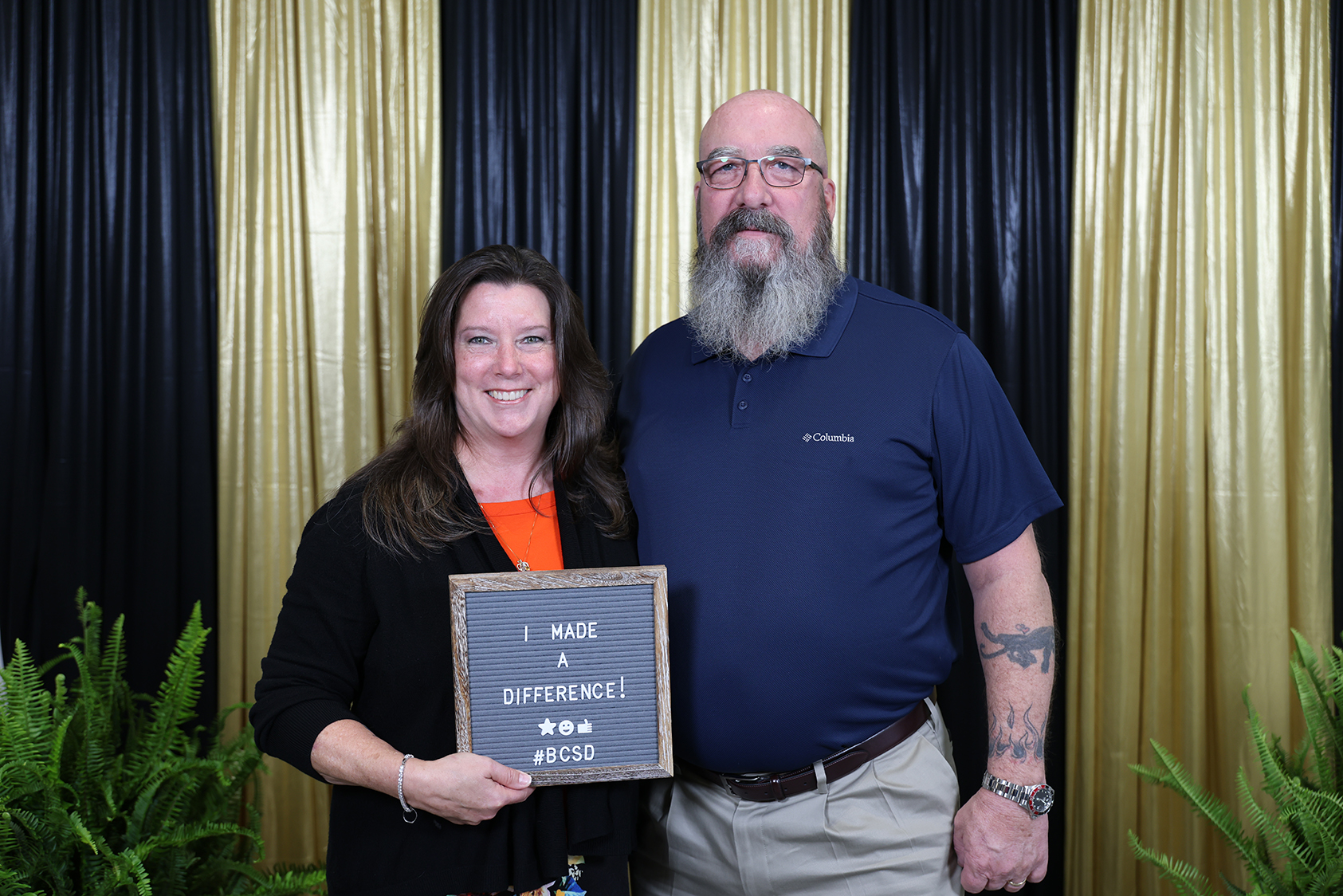A man and a woman stand together in front of a black and gold backdrop. The woman holds a sign.
