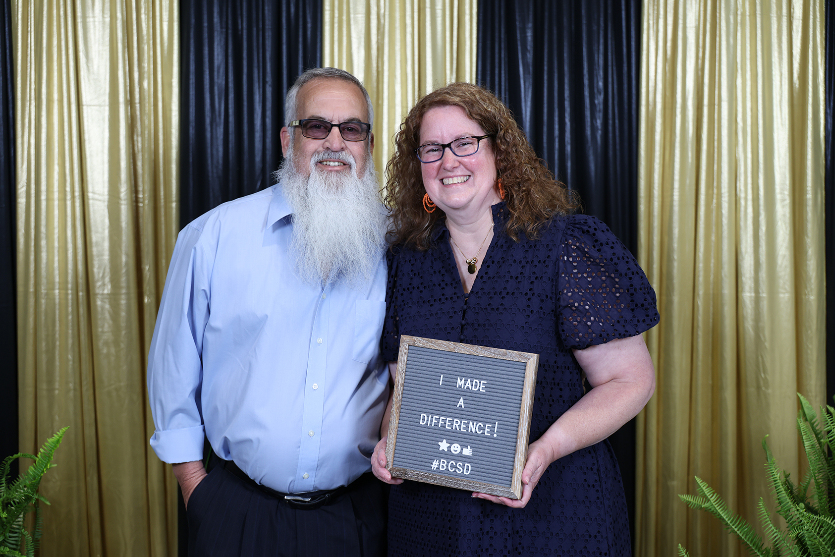 Two people pose for a picture with a plaque. A man with a long white beard and glasses stands beside a woman wearing glasses and a necklace.