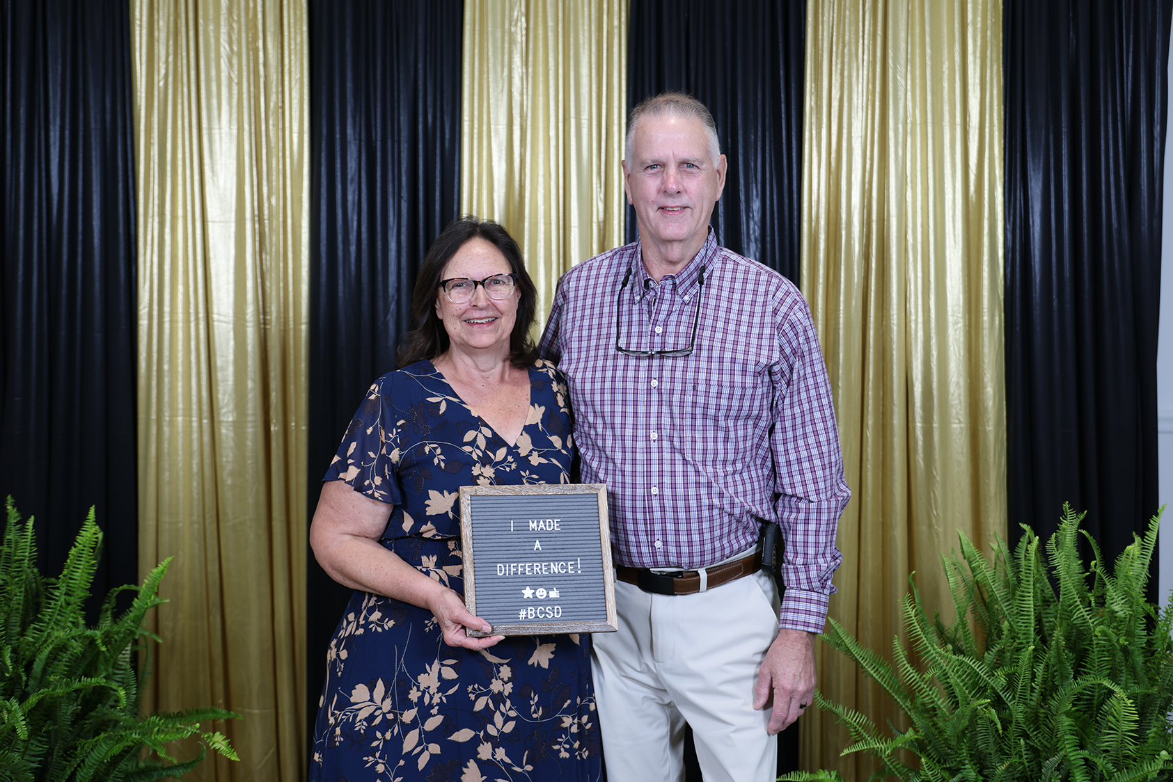 A man and a woman stand in front of a backdrop with gold and black drapes, holding a plaque.