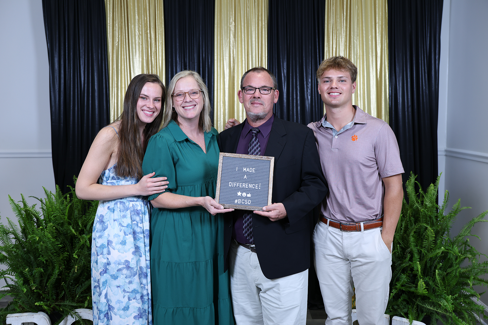 Four people standing together holding a plaque with text, posing in front of a black and gold curtain backdrop.
